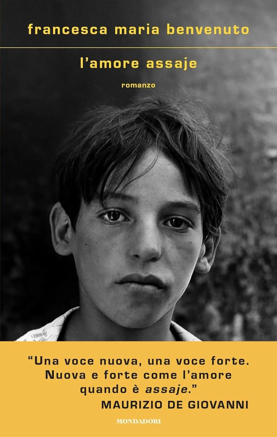 Book cover featuring a black-and-white photograph of a young boy with short hair, with yellow text in Italian, including the title "l'amore assaje" and a quote by Maurizio De Giovanni.