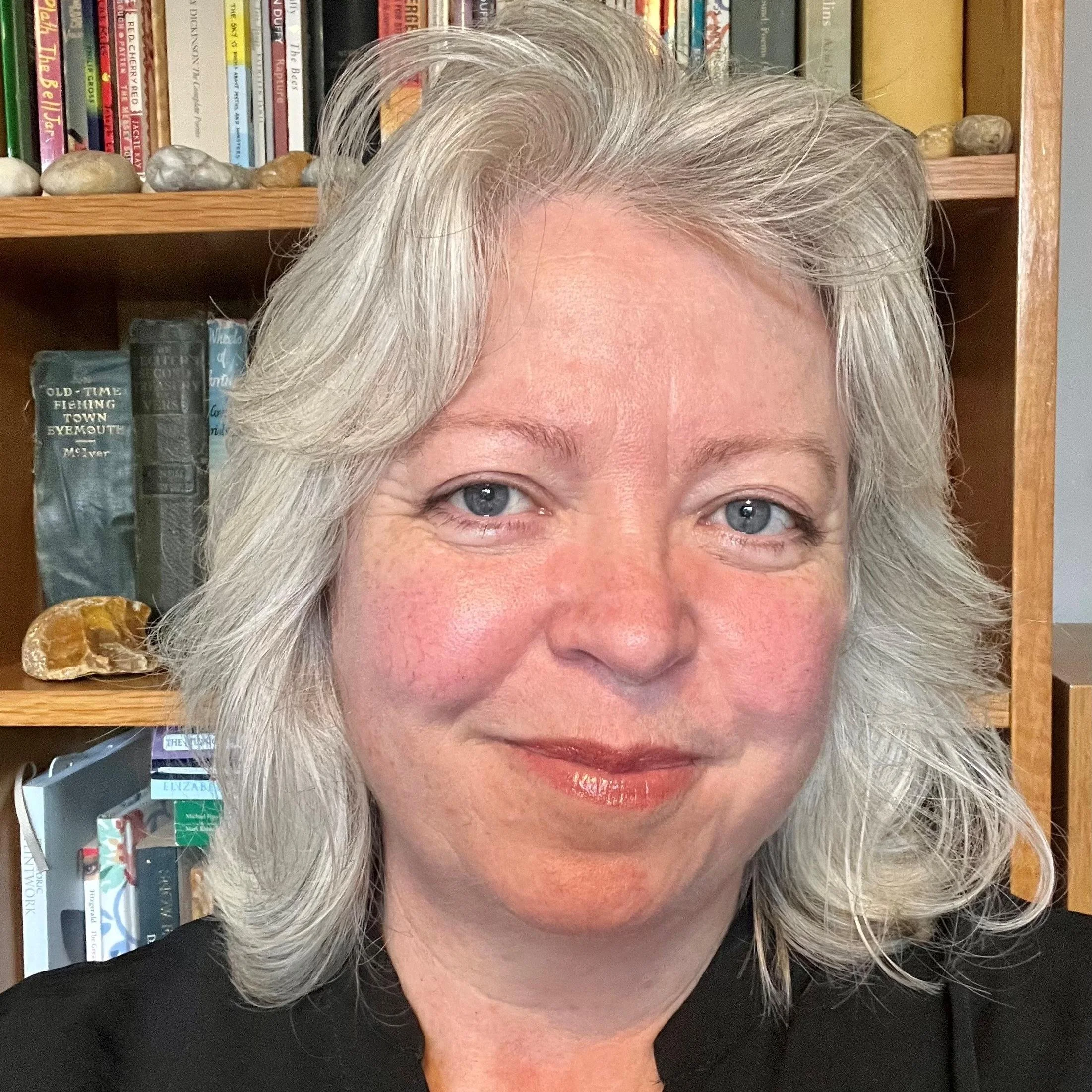 A middle-aged woman with gray hair and blue eyes smiling in front of a bookshelf filled with books and some decorative rocks.