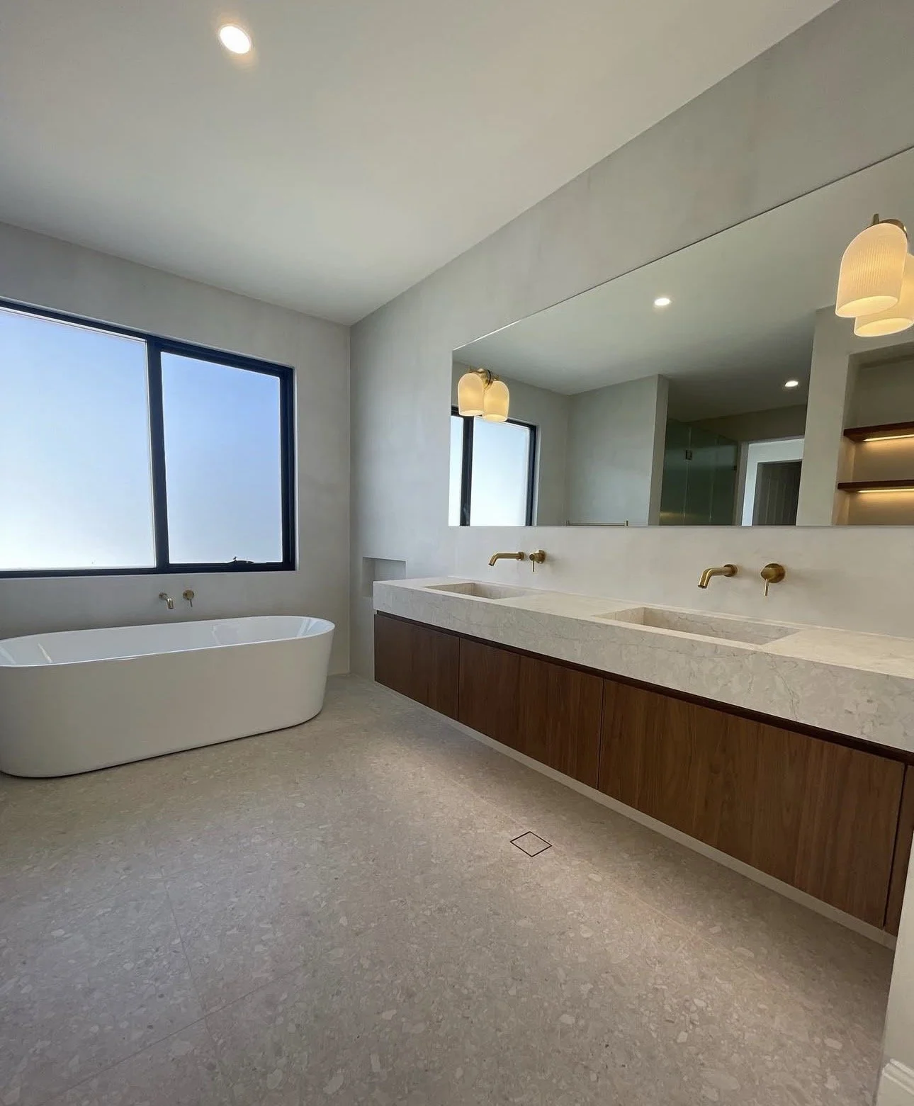Modern bathroom with large window, freestanding bathtub, double vanity with two sinks, large mirror, warm lighting, beige tile flooring, light-colored walls, minimal decor.