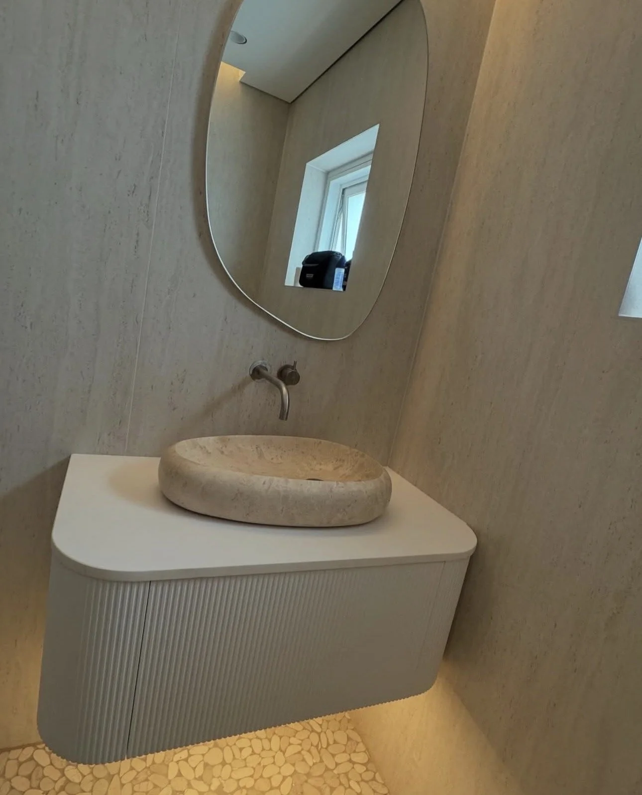 A wall-mounted bathroom sink with an oval-shaped stone basin, a round mirror above it, a small window with a camera on the windowsill, and a textured white cabinet under the sink.