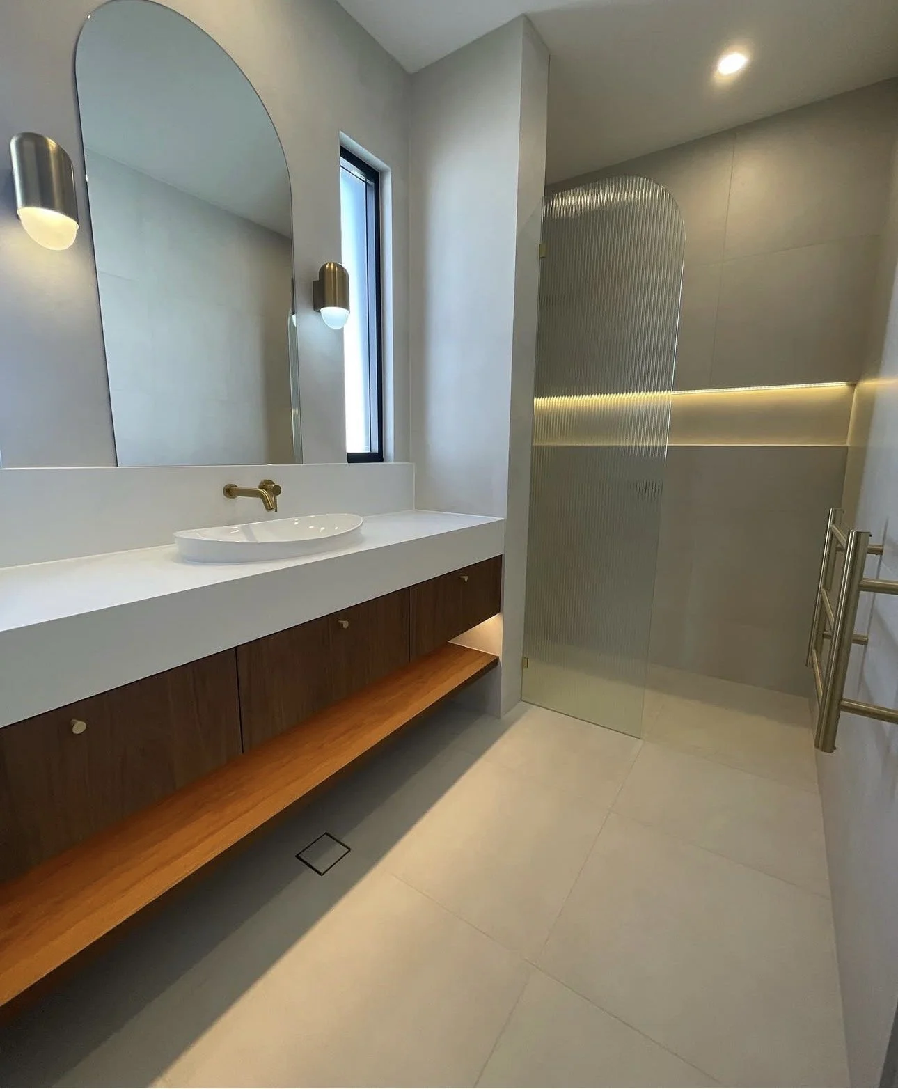 Modern bathroom with a white sink, wooden vanity, large mirror, wall-mounted light fixtures, narrow window, and a textured glass shower enclosure.