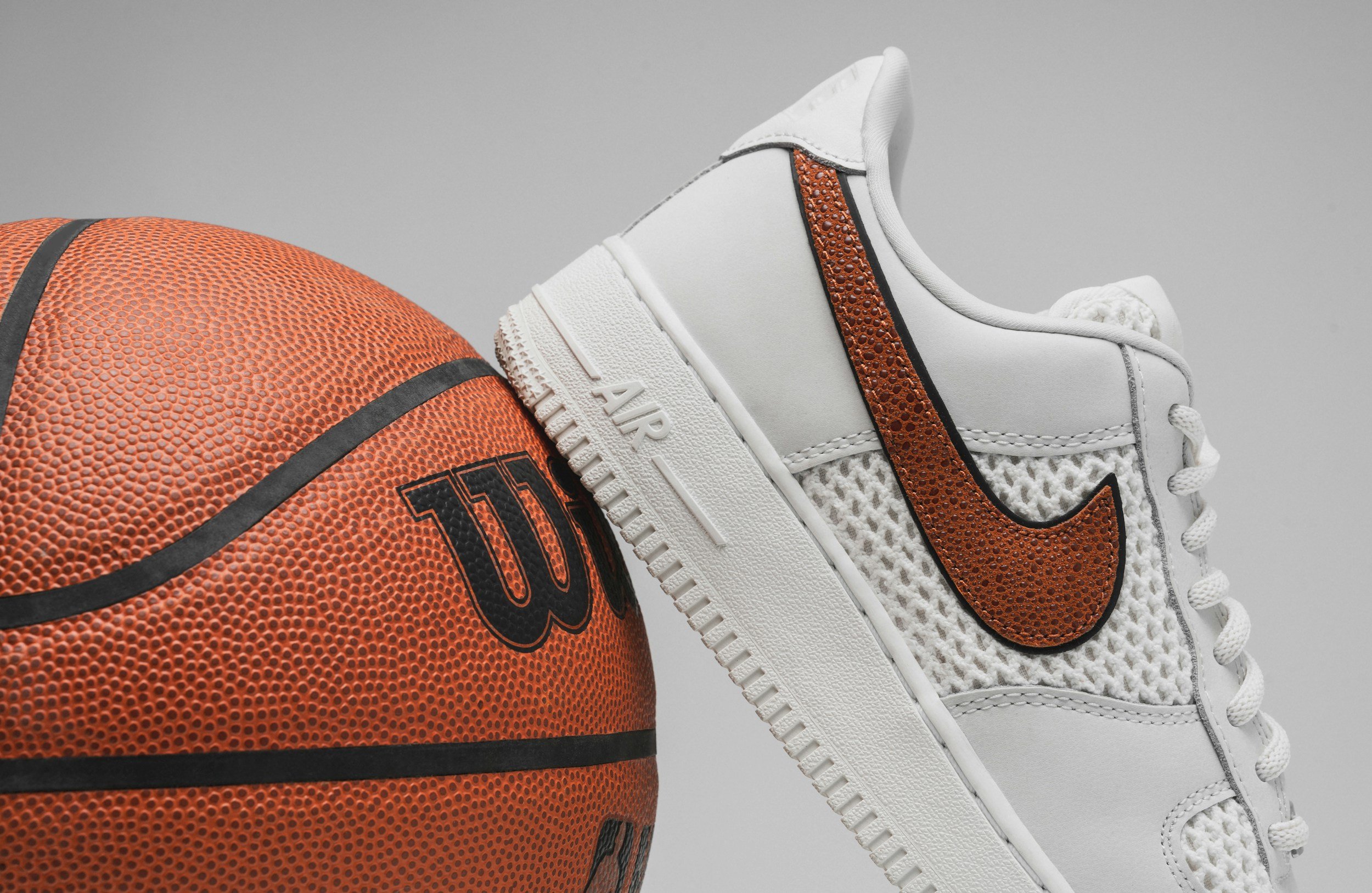 Close-up of a basketball and a white sneaker with a brown Nike swoosh logo and textured details against a plain gray background.