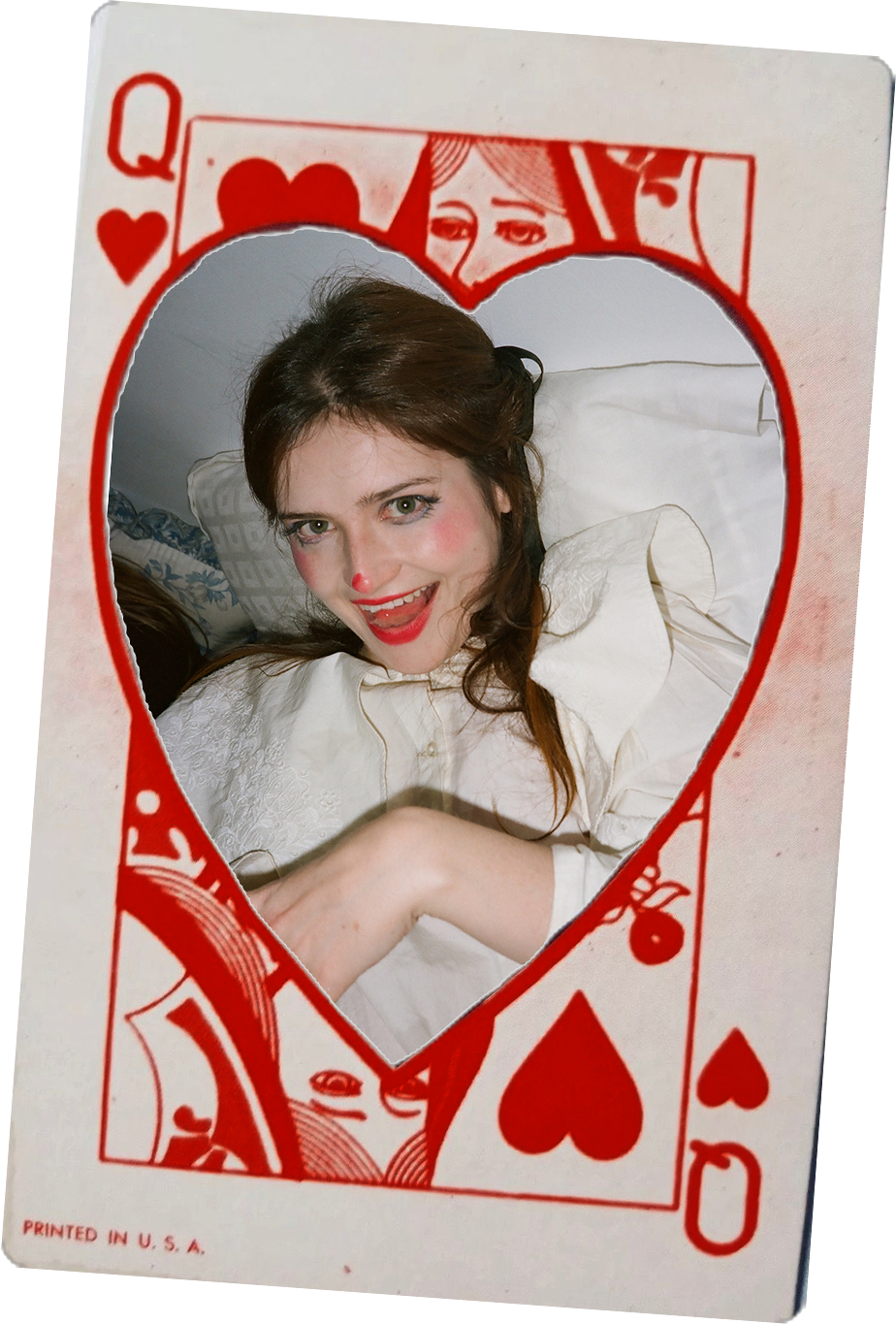 A woman with brown hair and fair skin dressed as a clown, with red lipstick, red nose, and blush, posing behind a cardboard cutout of a Queen of Hearts playing card with a heart-shaped window.