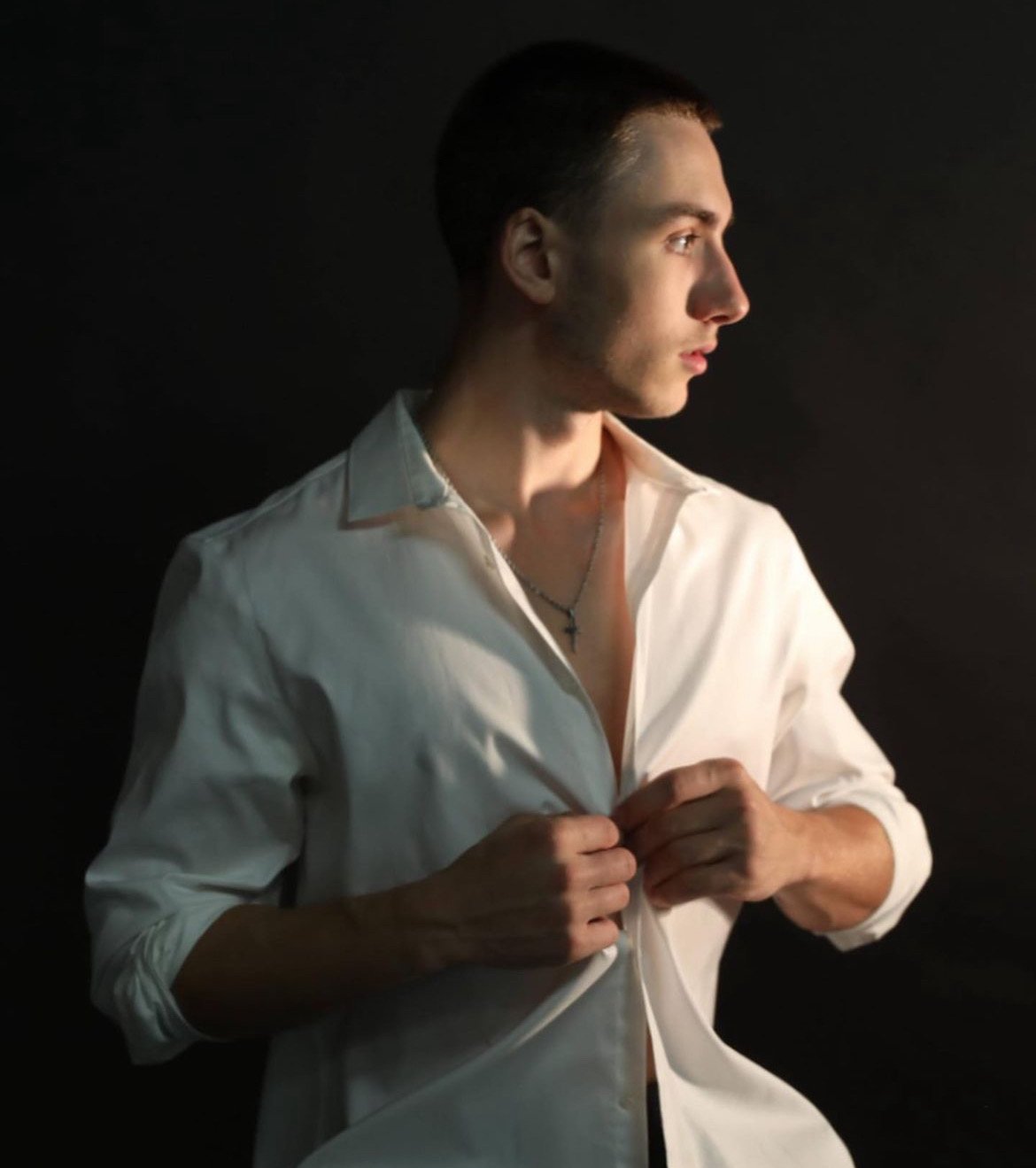 Ezra Urmy with short dark hair, wearing a partially unbuttoned white shirt, stands against a black background. He is looking to his right and is adjusting his shirt with both hands, wearing a cross necklace.