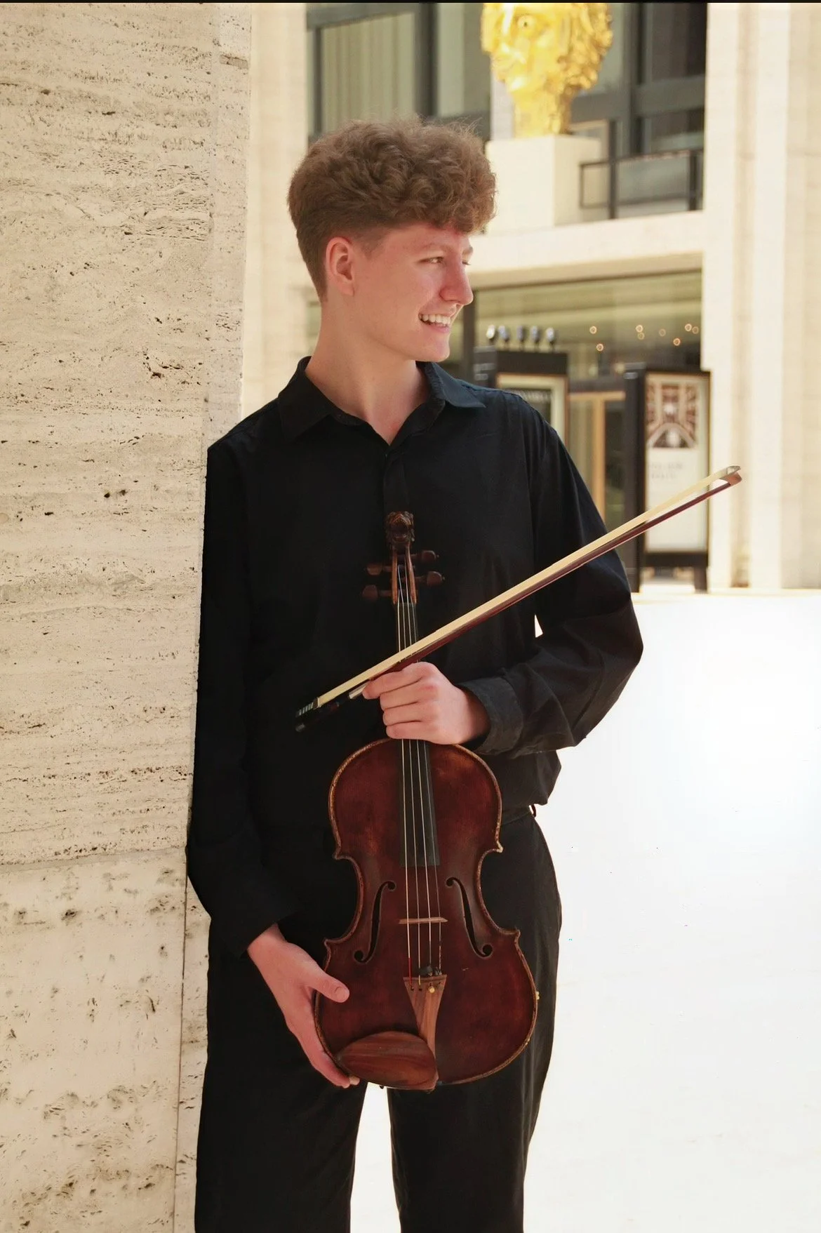 Young man with curly hair holding a violin and bow, standing against a building with large windows and light-colored stone walls, smiling and looking to the side.