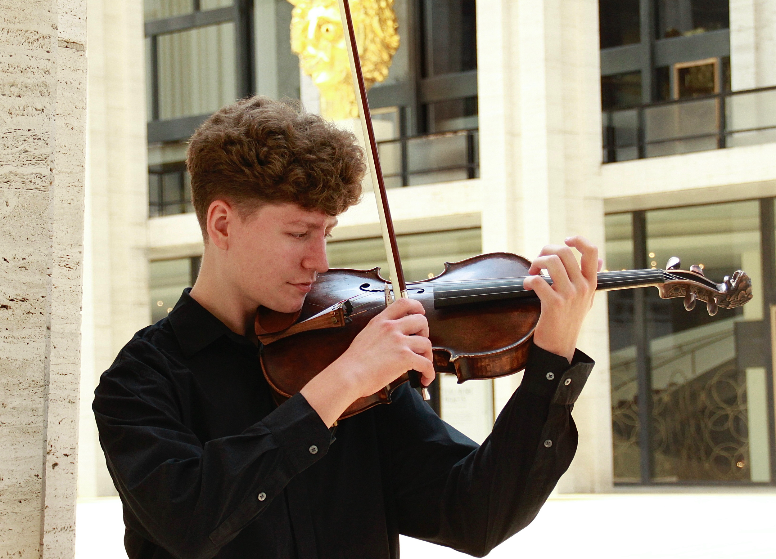 A young man with curly hair playing a violin outdoors, wearing a black shirt, with a modern building in the background.