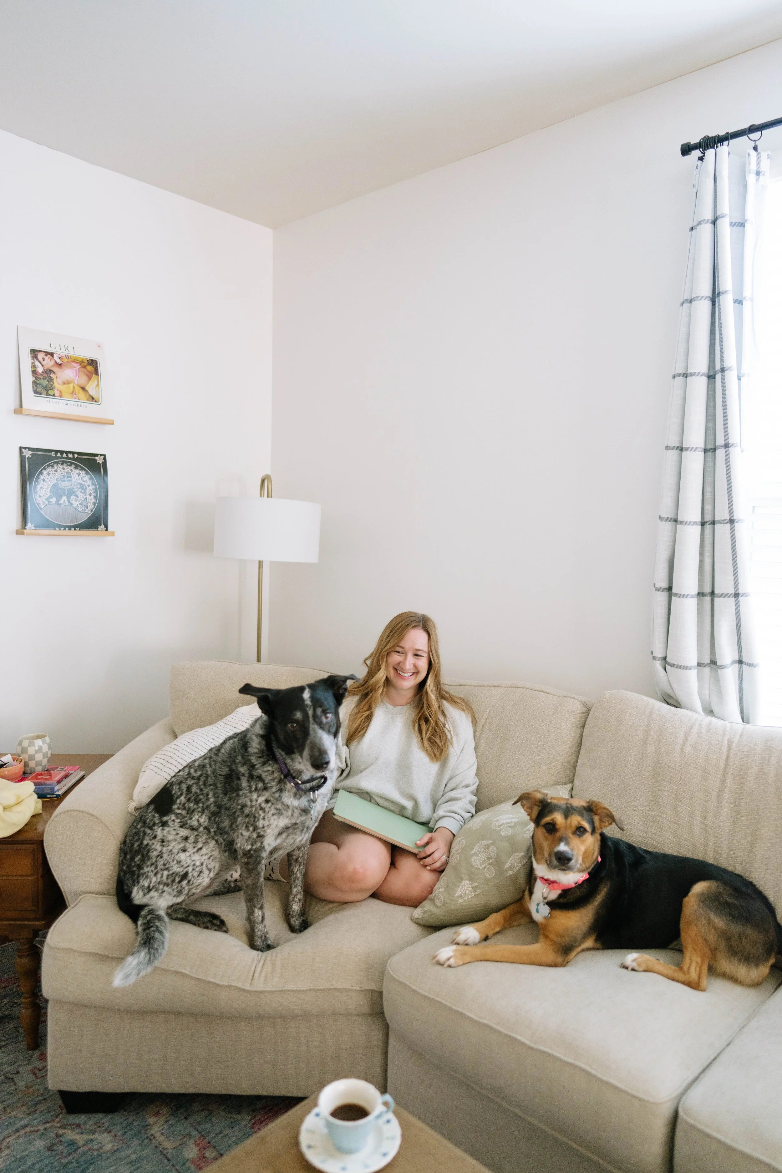 A young woman with long blonde hair sitting on a beige sofa in a bright living room, smiling, with three dogs around her. One large black and white dog is sitting on the sofa, another large black dog with tan markings and a red collar is lying on the sofa, and a smaller tan dog is resting on the sofa beside her. The room has white walls with two framed pictures, a floor lamp, and a window with white curtains. A coffee table with a cup of coffee and some other items is visible in the foreground.