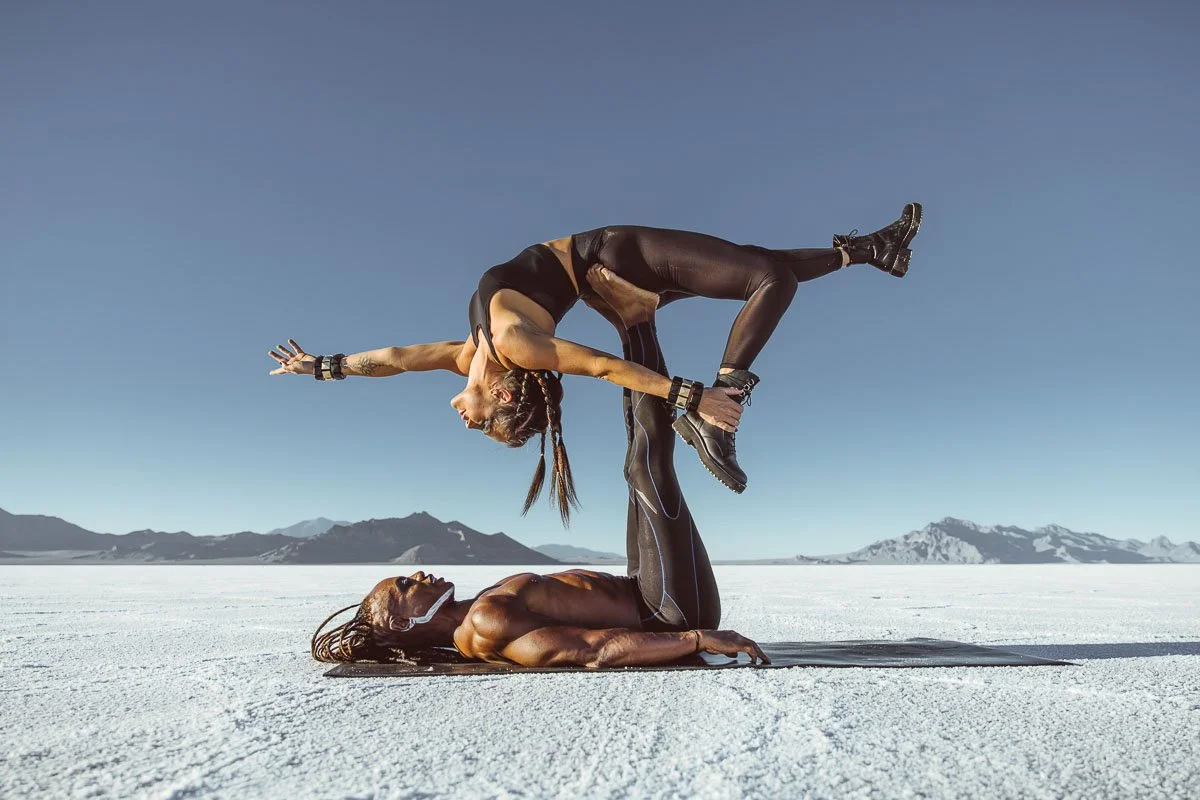 Two women practicing acro yoga outdoors on a dry salt flat, with mountains in the background. One woman is lying on the ground on her back, supporting the other woman who is balancing on her hands and feet in a pose.