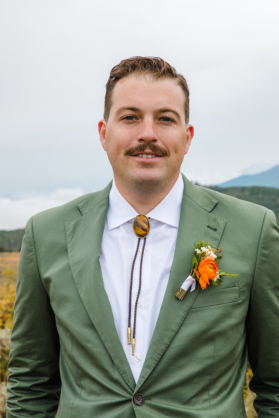 A man with a mustache wearing a green blazer, white shirt, and a bolo tie with a large amber stone, standing outdoors with a blurred landscape and mountains in the background.