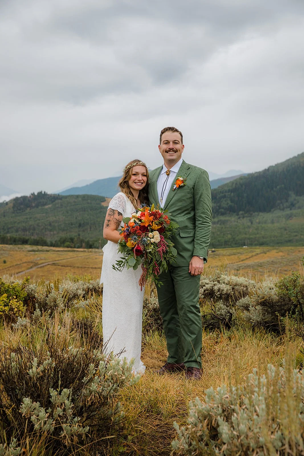 A bride and groom standing outdoors in a field with mountain views in the background, smiling at the camera, with the bride holding a large colorful bouquet.