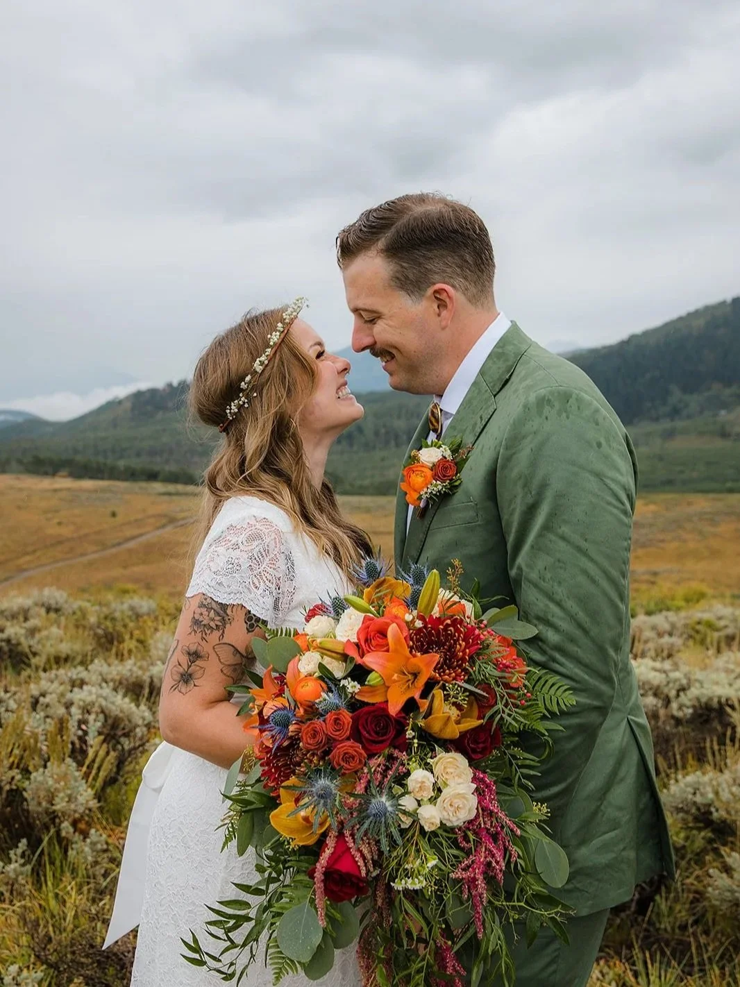 A bride and groom stand close, smiling and touching foreheads in a scenic outdoor landscape. The bride holds a large, colorful bouquet of flowers, and the groom is dressed in a green suit with a boutonniere. Overcast sky and rolling hills in the back