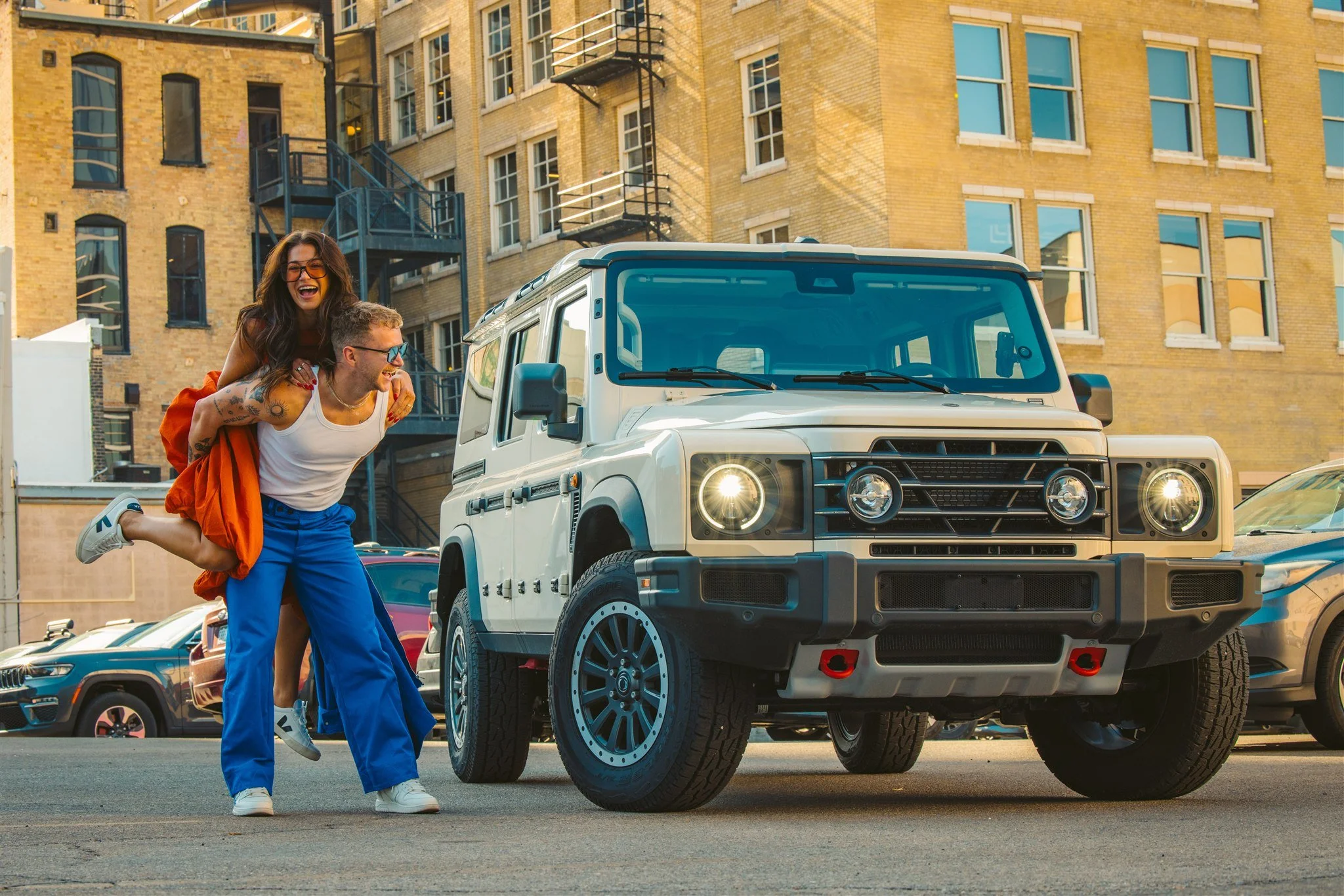 A woman giving a piggyback ride to a man next to a white off-road vehicle in an urban area with brick buildings and parked cars.
