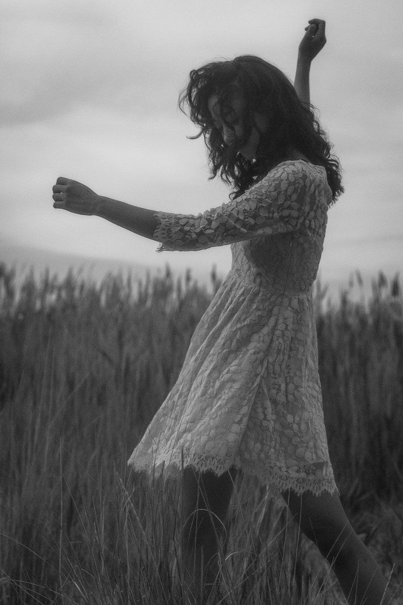 A woman with curly hair wearing a lace dress standing in a field, with her arms raised, in black and white.
