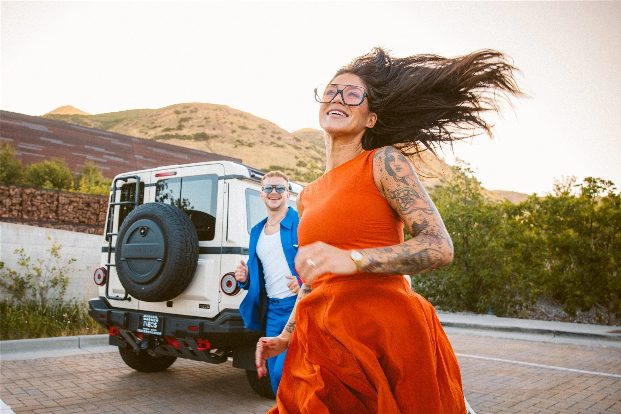 A woman with long hair, glasses, and tattoos on her arms, smiling and dancing outdoors in front of a white off-road vehicle. A man in sunglasses and a blue suit stands behind her, smiling. The background features a mountainous landscape and clear sky