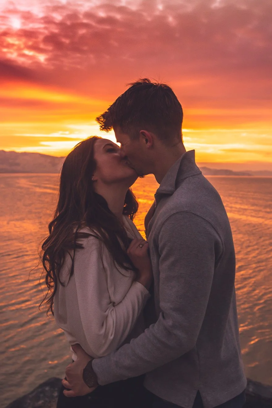 A couple kissing by the water at sunset with a vibrant orange and pink sky in the background.
