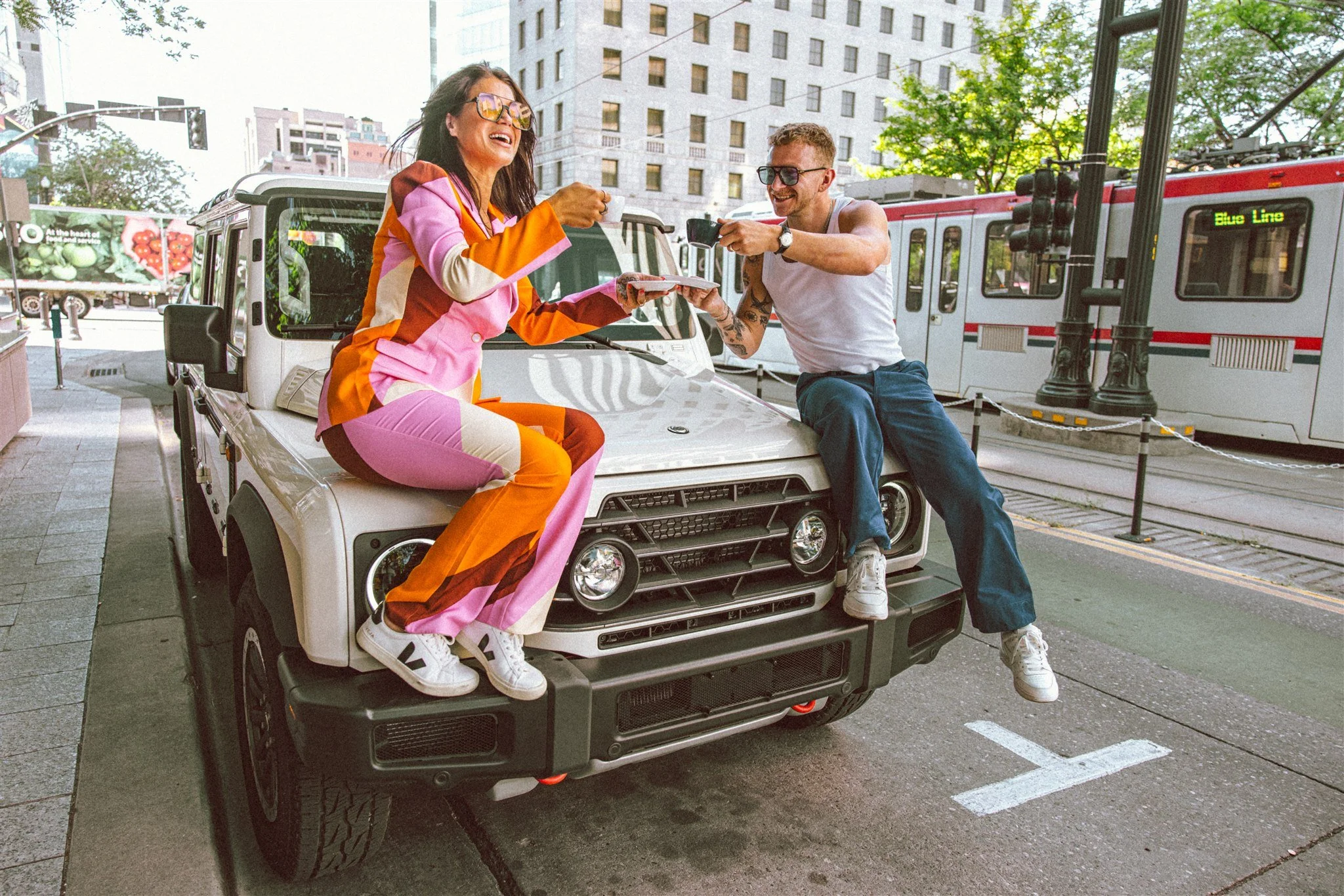A woman in a colorful, striped jumpsuit sitting on the hood of a white SUV, smiling and holding a mug, while a man in a white tank top and jeans, sitting on the SUV's hood, takes a photo of her with a camera. They are outdoors on a city street with a
