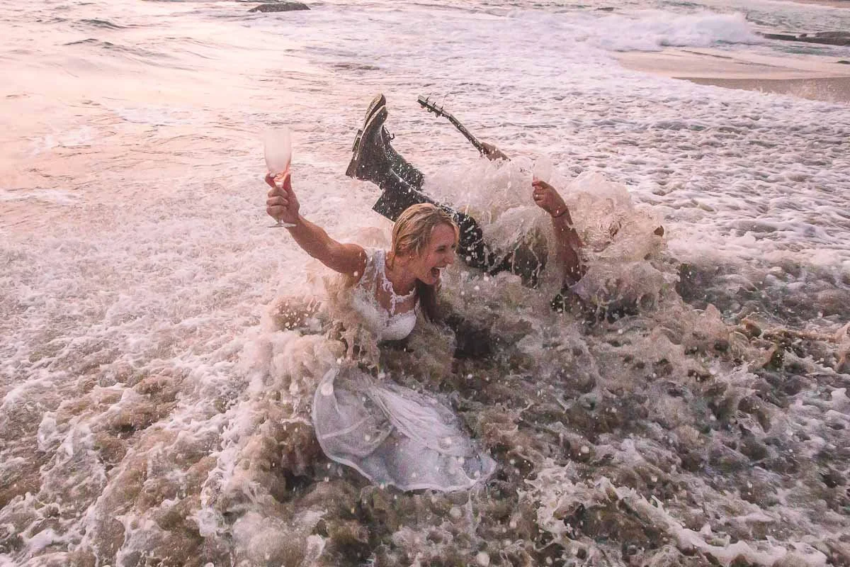 A woman in a white dress falling into the water at the beach while holding a glass of wine in each hand.