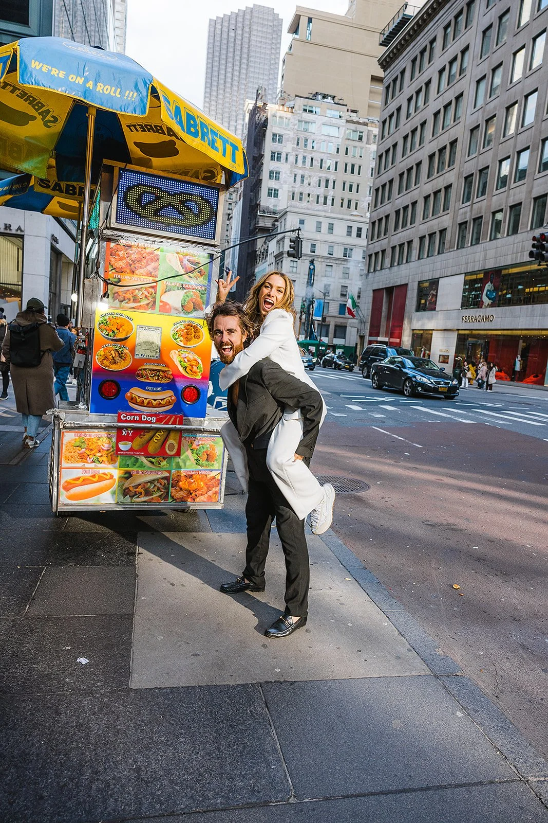 Two happy people, a man and a woman, are posing playfully on a city street with food cart. The man is carrying the woman on his back, both smiling and making playful gestures. The background shows tall buildings, cars, and pedestrians in an urban sho