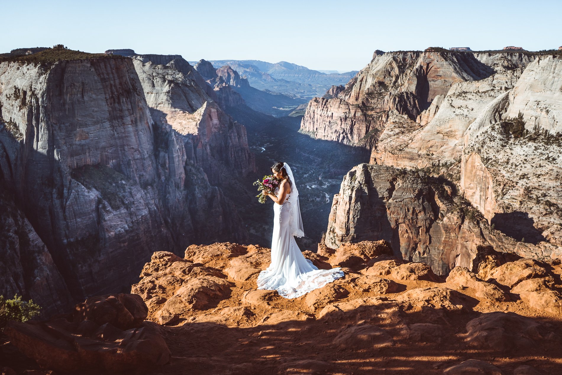 A bride in a white wedding dress holding a bouquet of flowers stands on a rocky ledge at a scenic canyon overlook with dramatic cliffs and distant mountains.