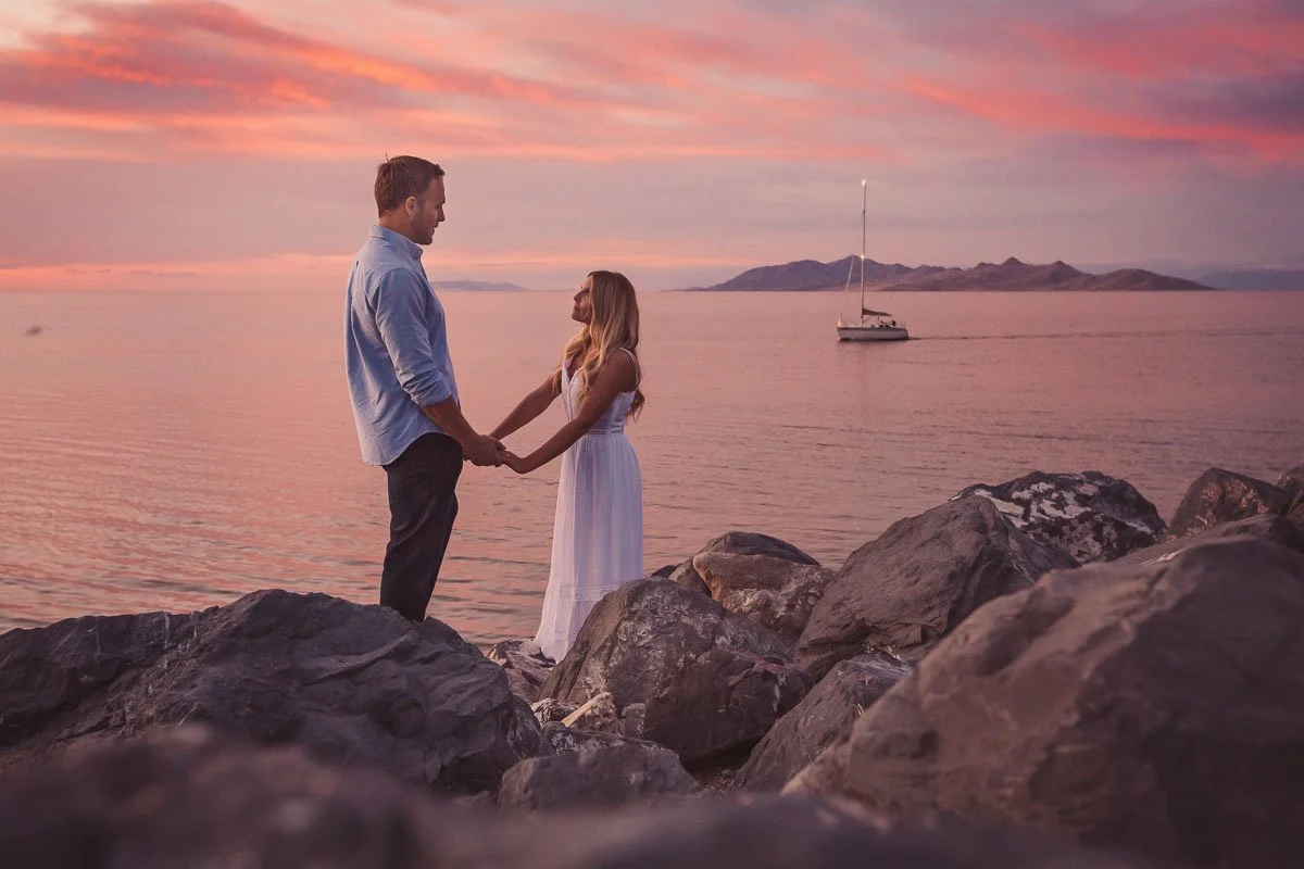 A couple holding hands standing on rocks by the water during a sunset with a sailboat in the background.
