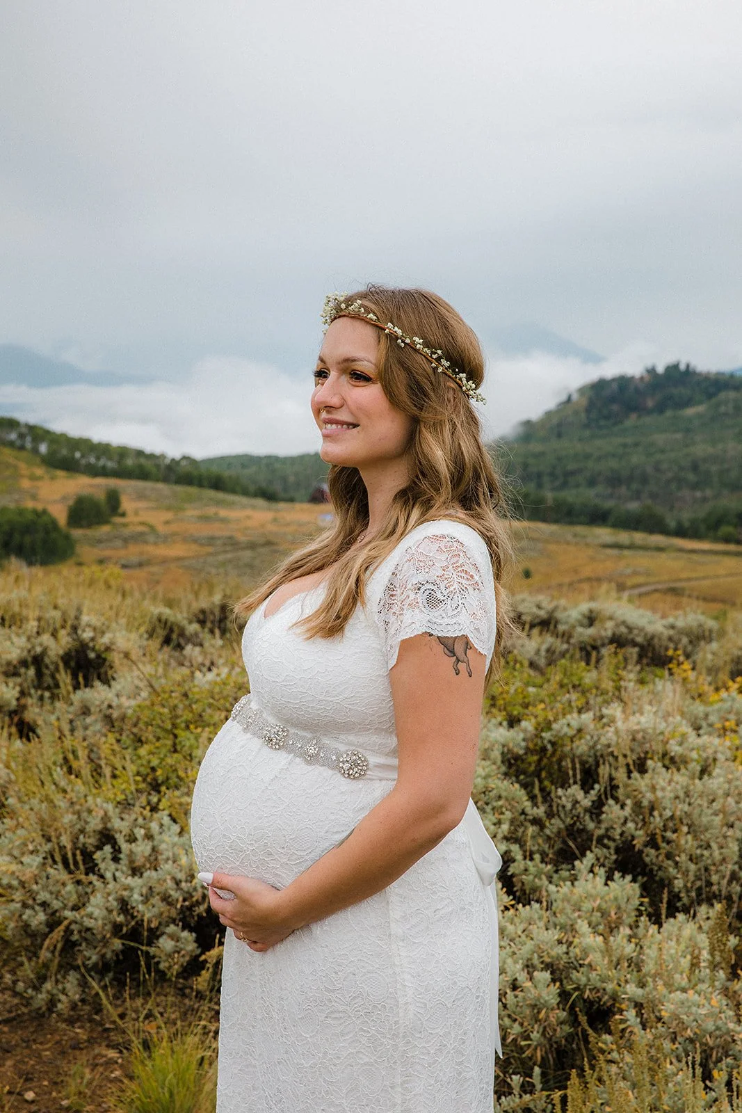 A pregnant woman wearing a white lace dress with a decorative belt, a floral headband, and a tattoo on her right arm, standing outdoors with a scenic landscape of rolling hills and greenery in the background.