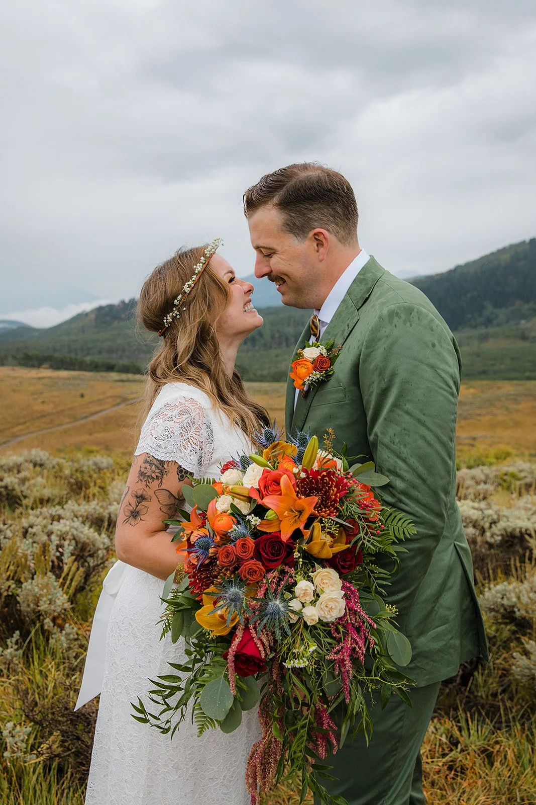 A couple in wedding attire touching foreheads outdoors on a cloudy day, with a woman holding a large colorful bouquet and a scenic landscape of hills and fields in the background.