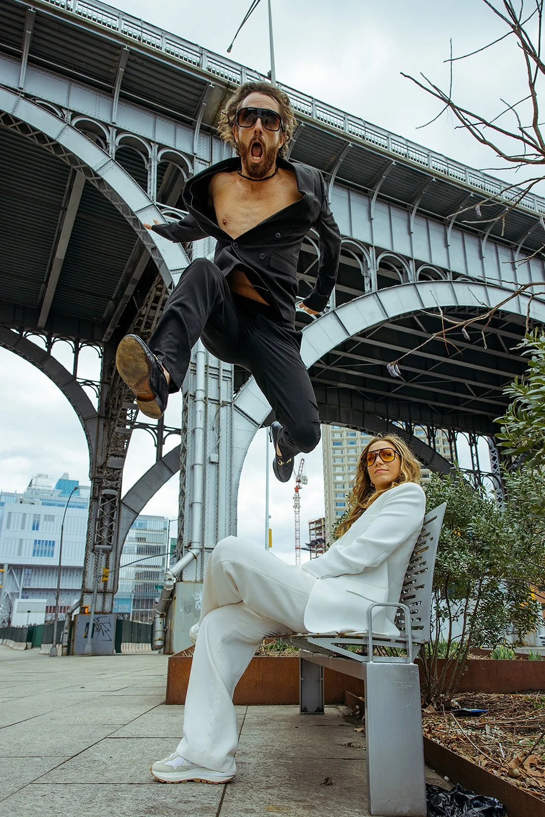 A man in a black suit is jumping in mid-air with his mouth open wide, and a woman in a white suit with sunglasses is sitting on a bench underneath, with an urban bridge and city buildings in the background.