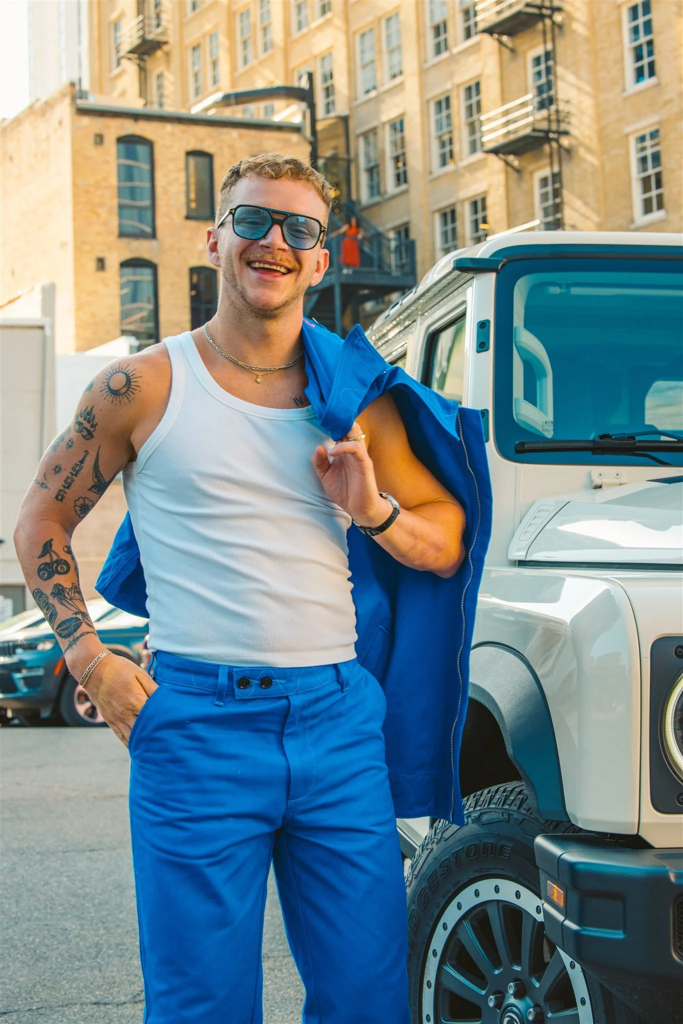 A young man with tattoos, wearing sunglasses, a white tank top, and blue pants, smiling and holding a blue jacket over his shoulder, standing beside a white vehicle on an urban street with buildings in the background.
