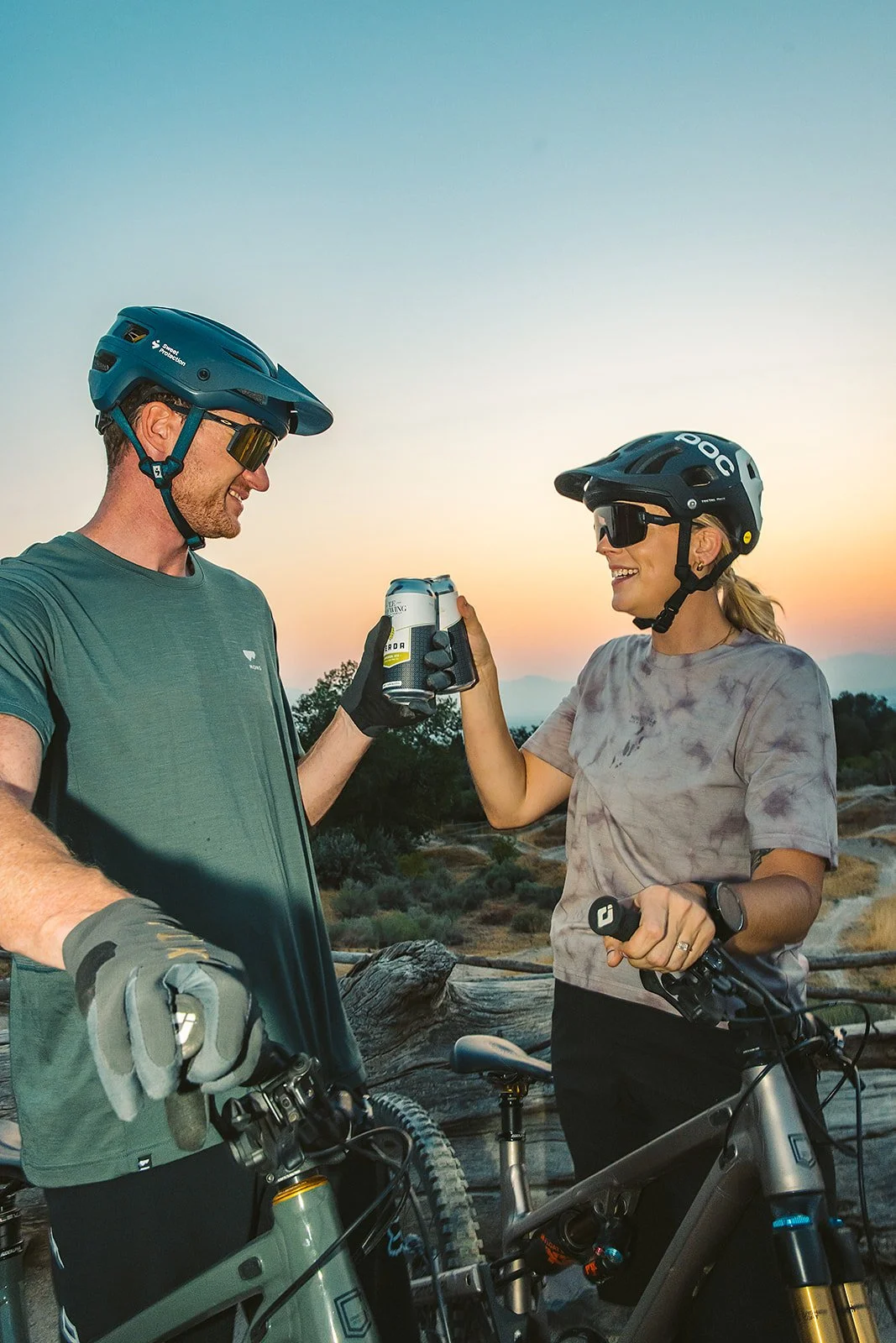 A man and woman in mountain biking gear sharing a drink at sunset in a desert landscape.
