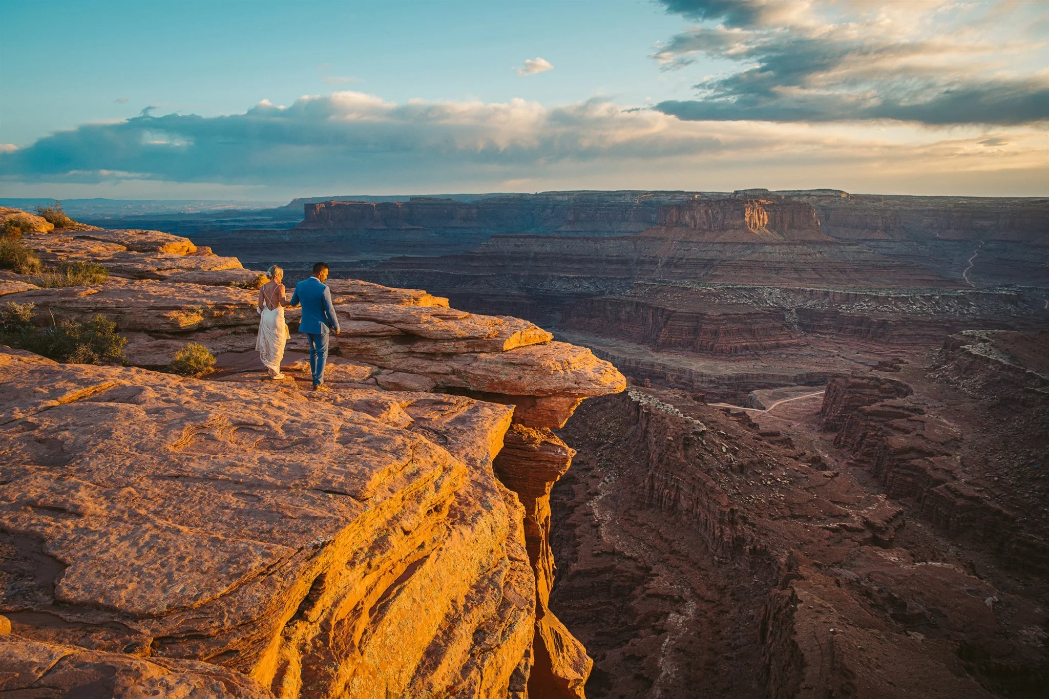 A couple dressed in formal attire walking along the edge of a rocky canyon during sunset.