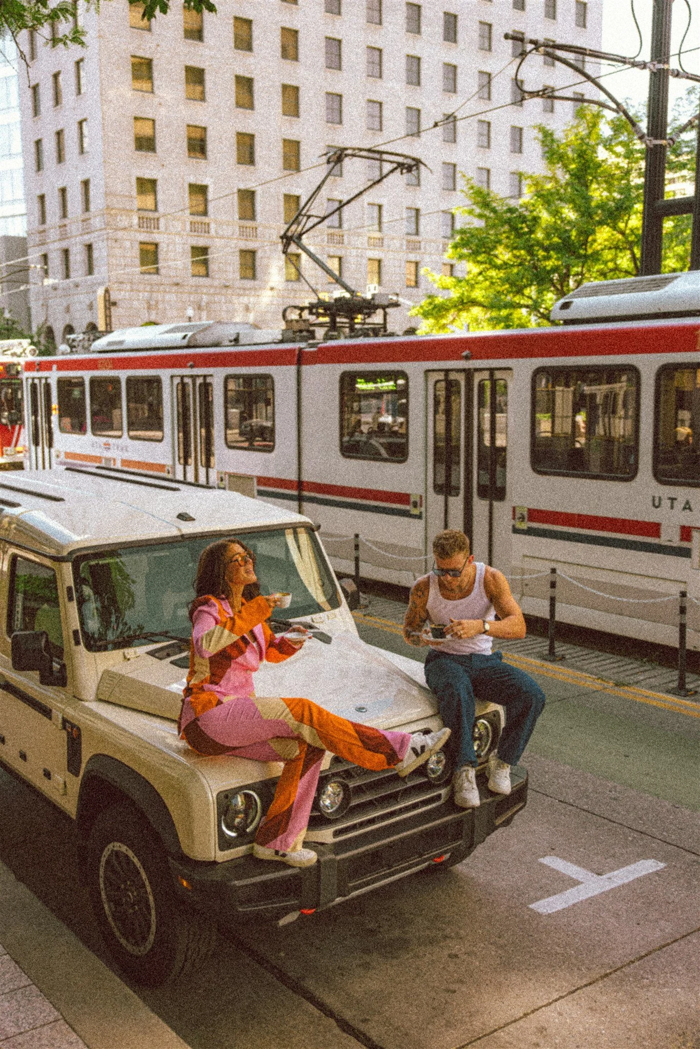 Two people sitting on the front of a beige Land Rover parked on a city street, with a light rail train behind them. The woman is wearing a colorful, patchwork jumpsuit and eating, while the man is in a white tank top and jewelry, looking at his phone
