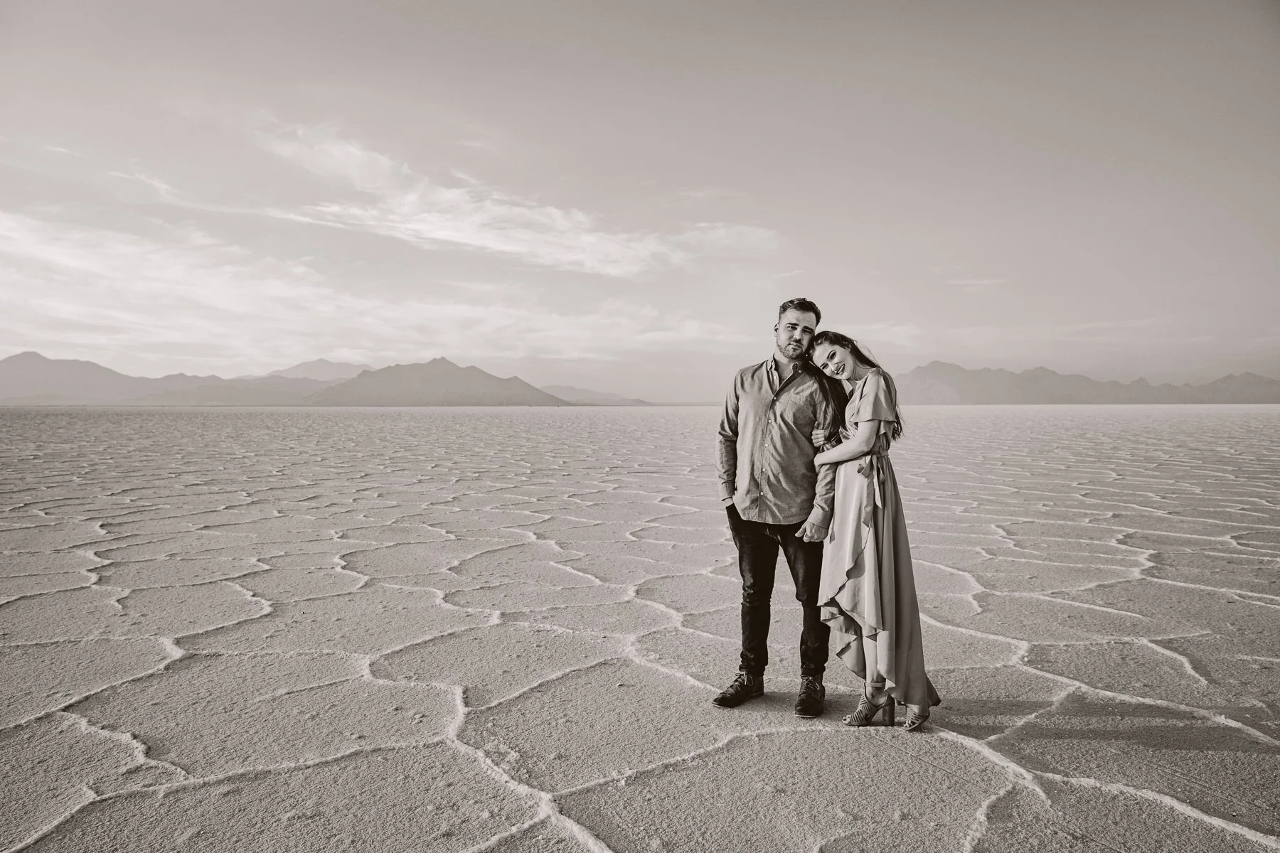 A black and white photo of a couple standing on a cracked desert landscape with mountains in the background, the woman resting her head on the man's shoulder.