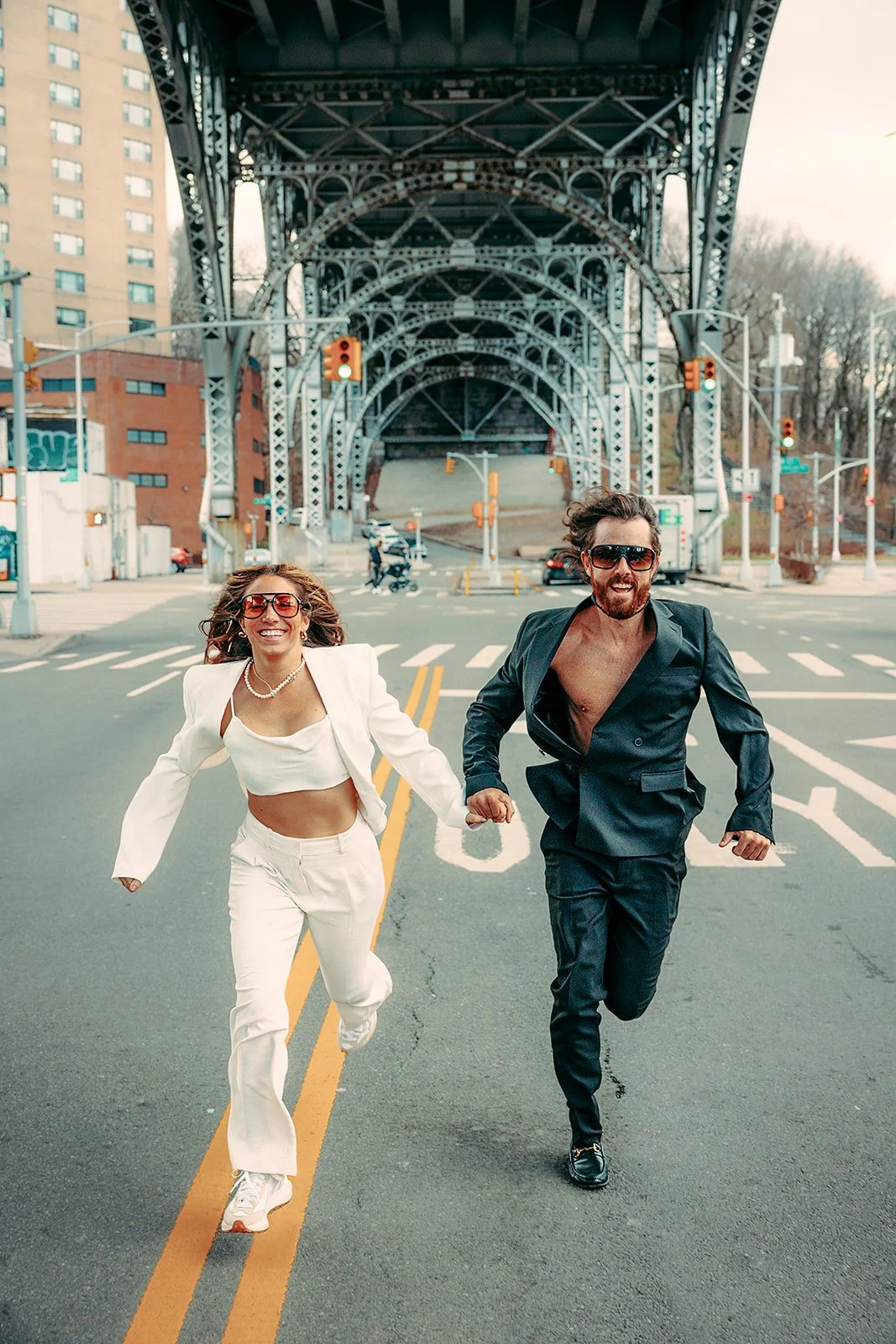 A man and woman in stylish outfits running across an empty city street under a large bridge, with skyscrapers and traffic signals in the background.