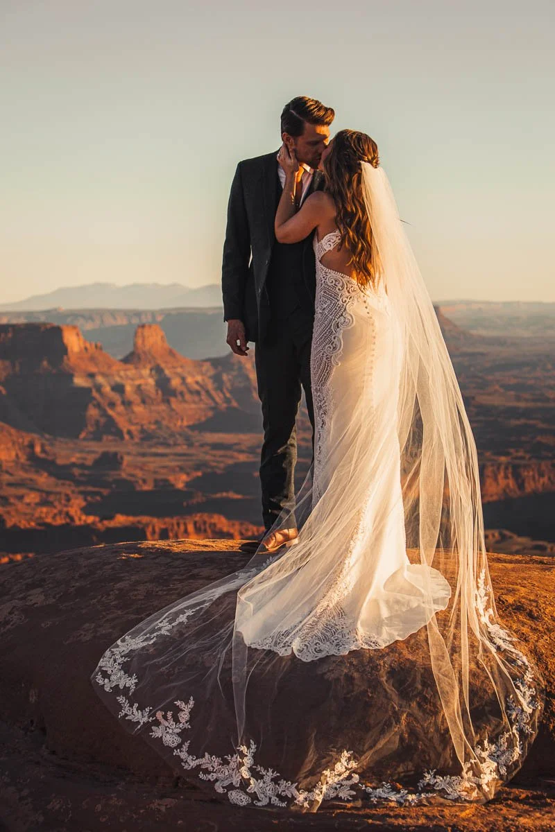 A bride and groom embracing on a rock with a desert canyon landscape in the background, during sunset.