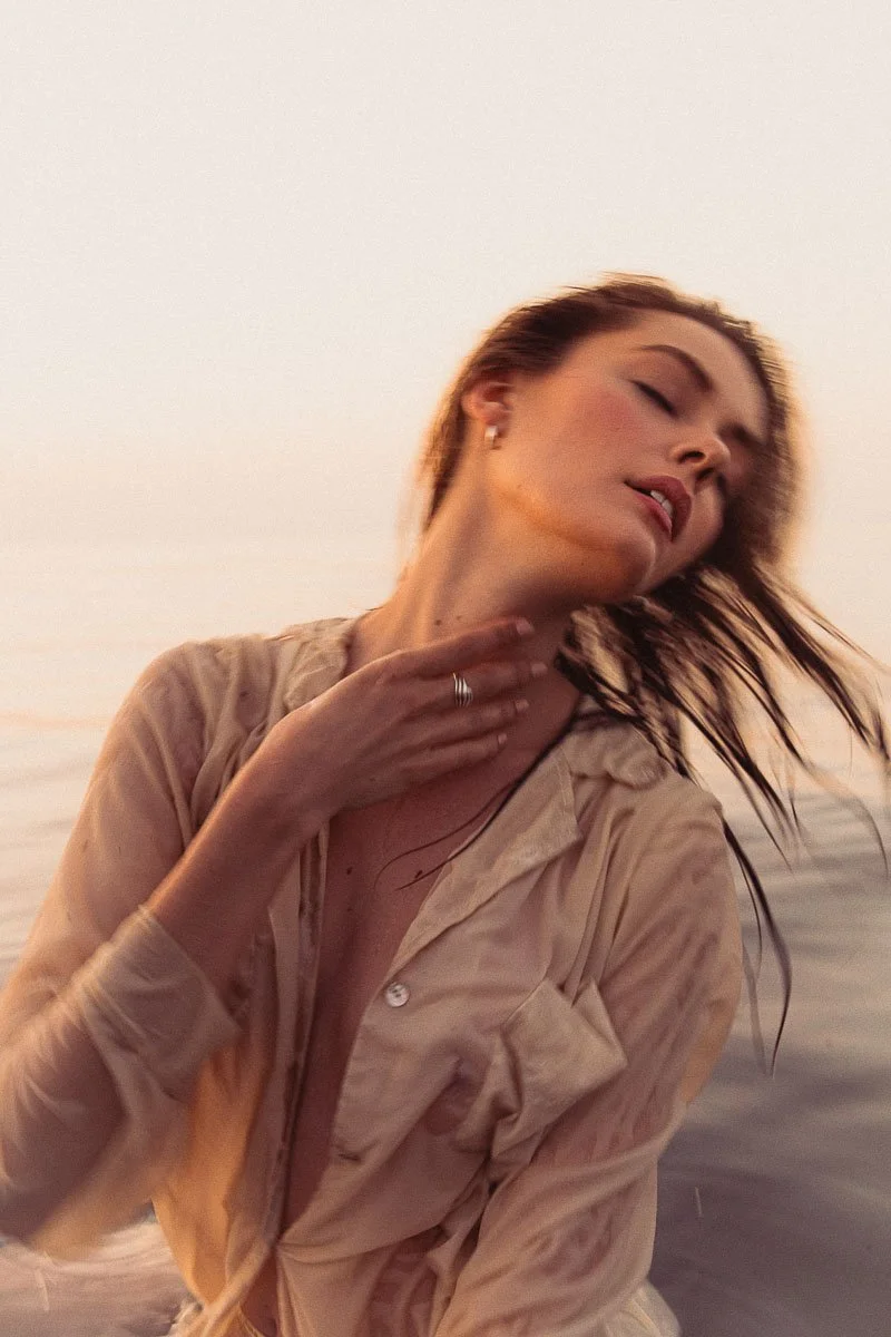 A woman with wet hair and a beige shirt tilting her head back with her eyes closed, near a body of water at sunset.