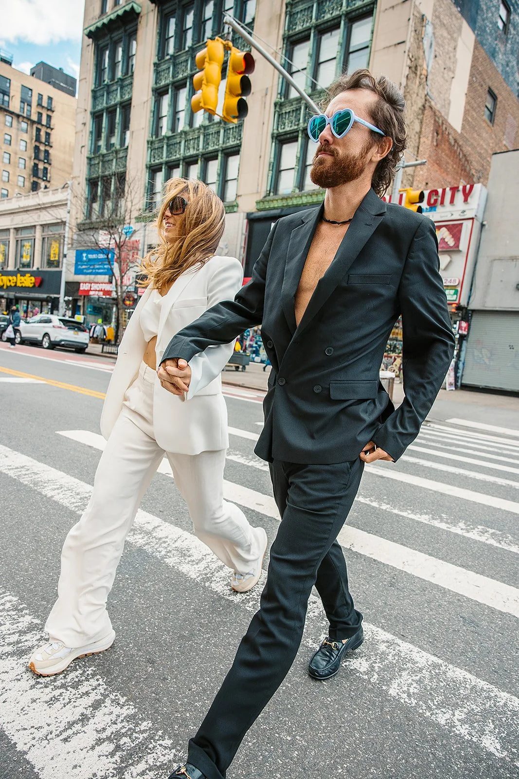 Two young adults, a man and a woman, holding hands while crossing a city street. The man has a beard, wears sunglasses with heart-shaped frames, a black suit with no shirt underneath, and black shoes. The woman has wavy hair, wears sunglasses, a whit