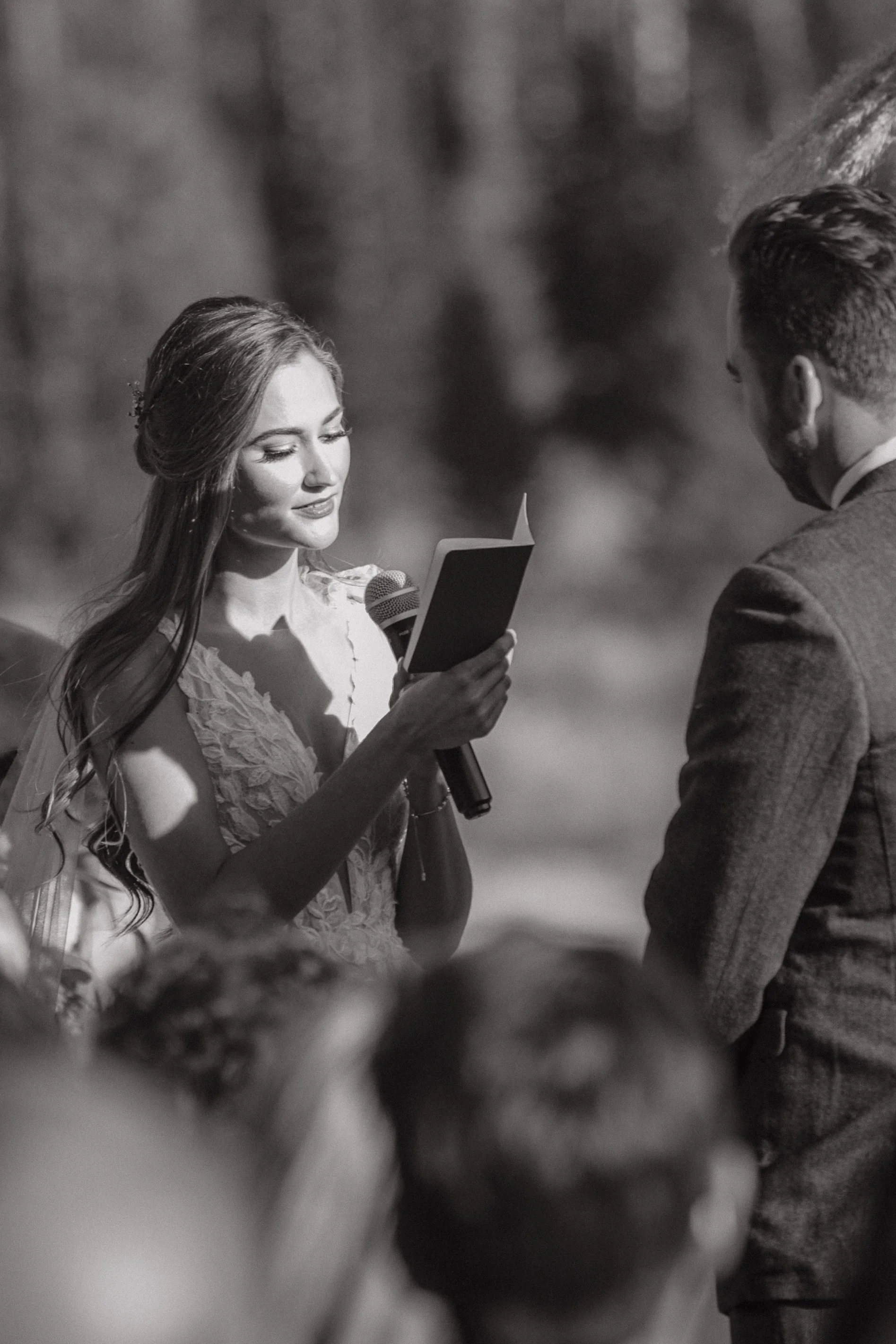 A woman in a wedding dress reading her vows to a man in a suit, outdoors, during a wedding ceremony.