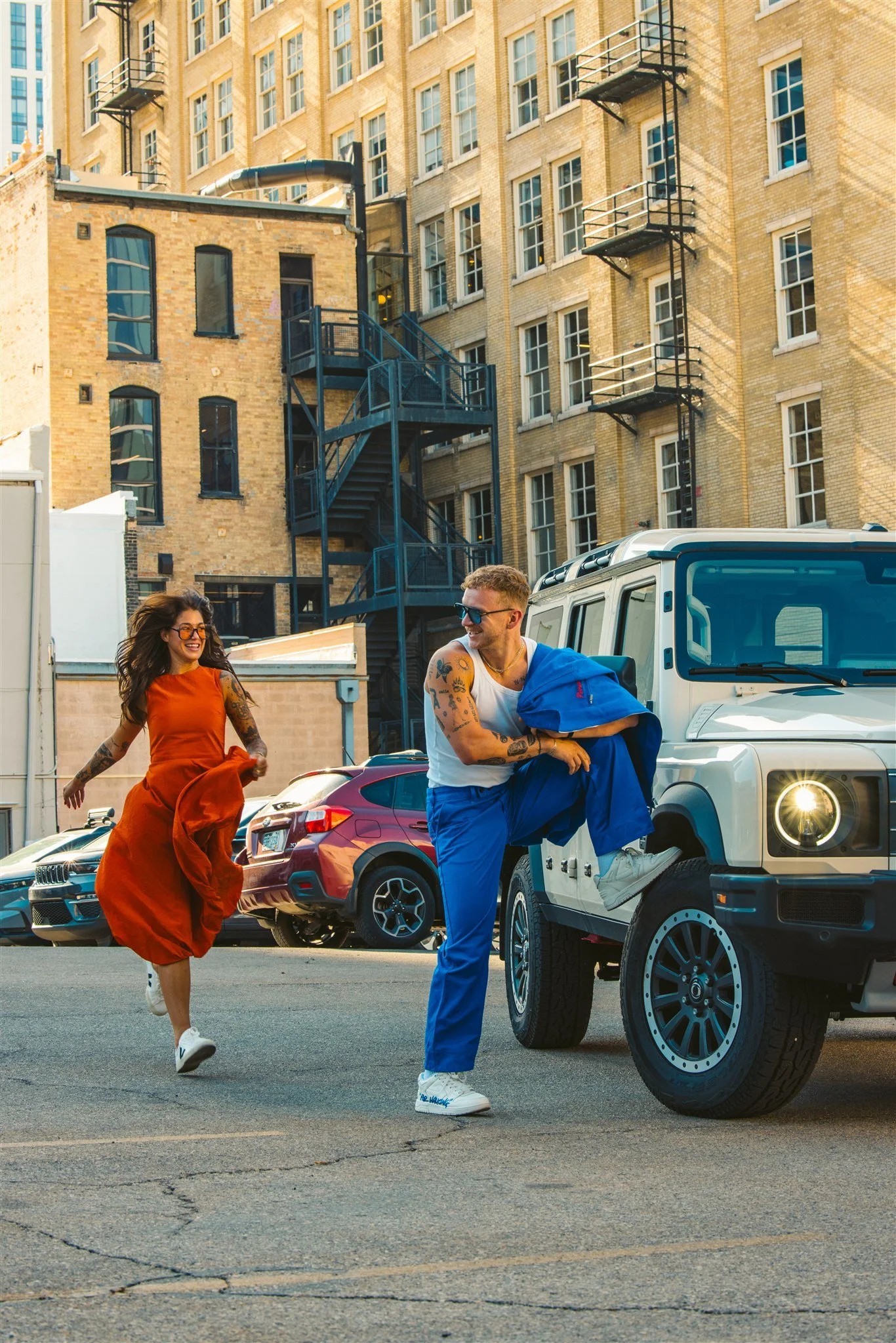 A man and woman enjoying themselves in an urban setting with parked cars and brick buildings in the background. The woman is running happily while the man is leaning on a white vehicle and laughing.