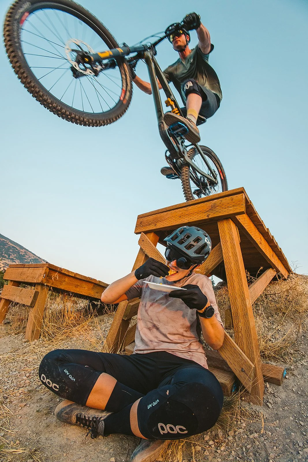 A mountain biker performs a stunt on a ramp in an outdoor setting with dry grass and mountains in the background. A person sitting beneath the ramp, wearing a helmet and gloves, is eating a snack.