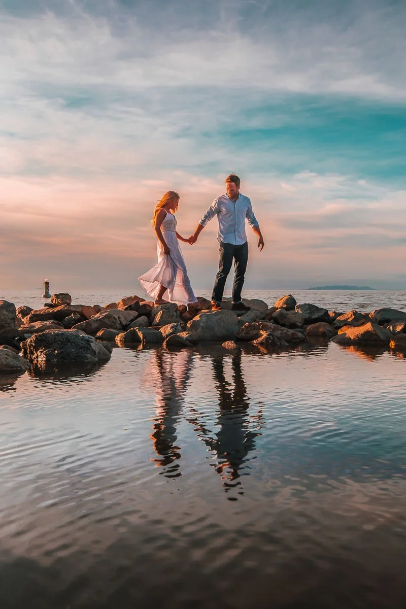 A couple holding hands and walking on rocks at the beach during sunset, with their reflections visible in the water.