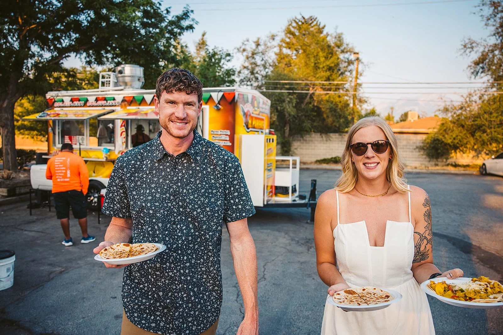 A man and woman holding plates of food, standing outdoors near a colorful food truck, with a man in an orange shirt preparing food in the background, trees, and parked cars.