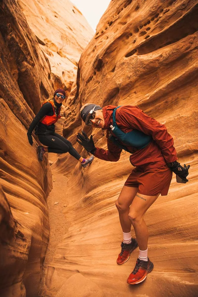 Two hikers smiling and giving high-fives in a narrow sandstone canyon with smooth, layered rock formations.