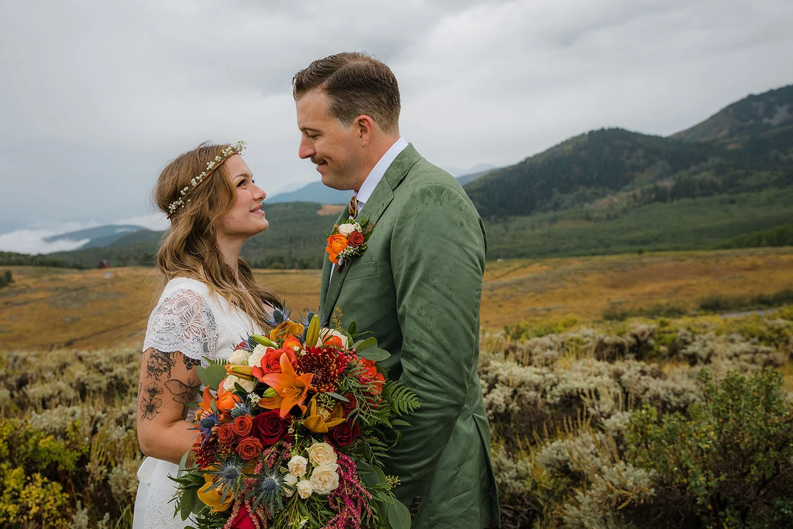 A bride and groom standing in a scenic outdoor landscape, gazing into each other's eyes, with mountains and cloudy sky in the background. The bride has tattoos on her arm and is holding a large colorful bouquet of flowers, while the groom is wearing 