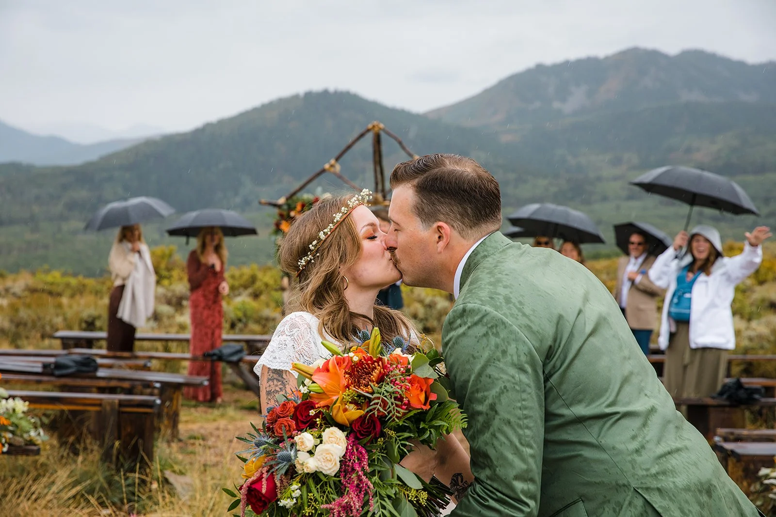 A couple kiss during their outdoor wedding in a mountainous landscape, with the bride holding a large colorful bouquet and surrounded by guests holding black umbrellas in rainy weather.