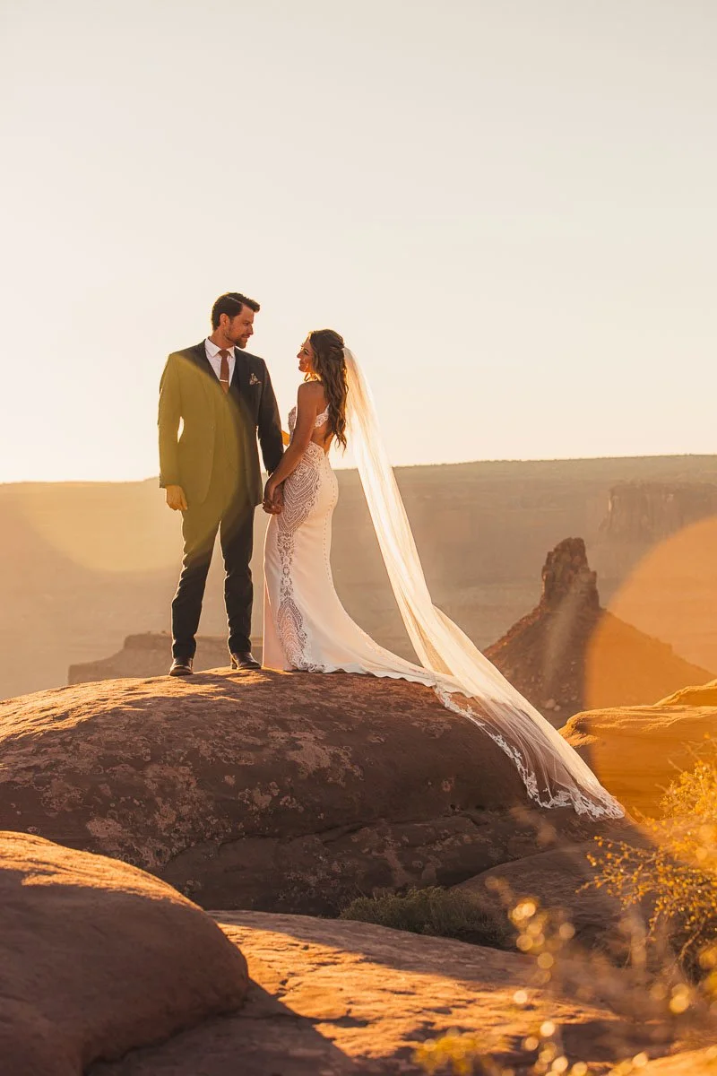 A bride and groom holding hands on a rocky landscape at sunset, with a distant rock formation and warm golden lighting.