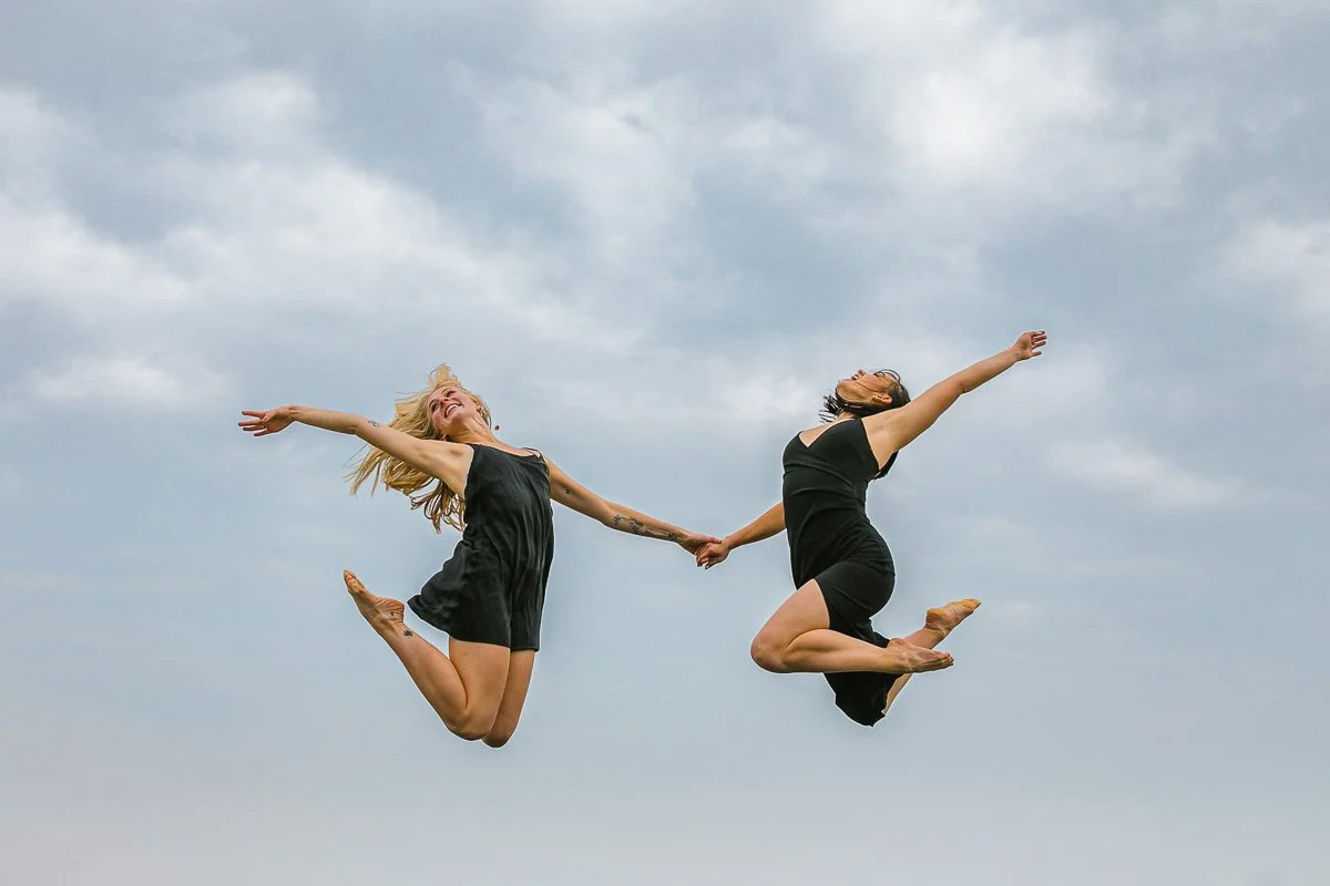 Two women wearing black dresses jumping in the air holding hands against a cloudy sky.