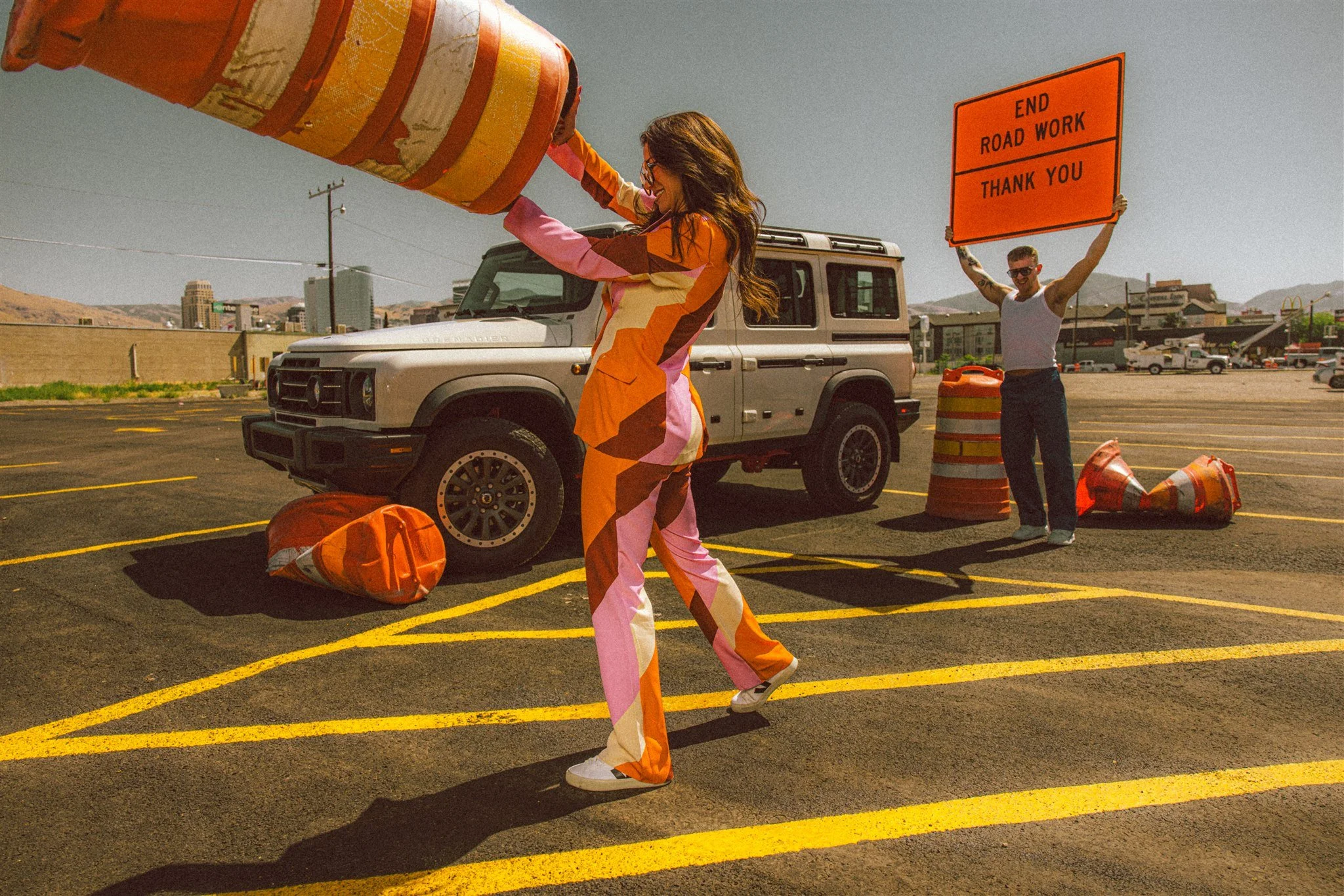 A woman in colorful pants and a patterned jacket is pulling away orange traffic cones in a parking lot. A man behind her is holding a large orange sign that reads 'END ROAD WORK THANK YOU.' There is a white off-road vehicle and traffic barrels in the