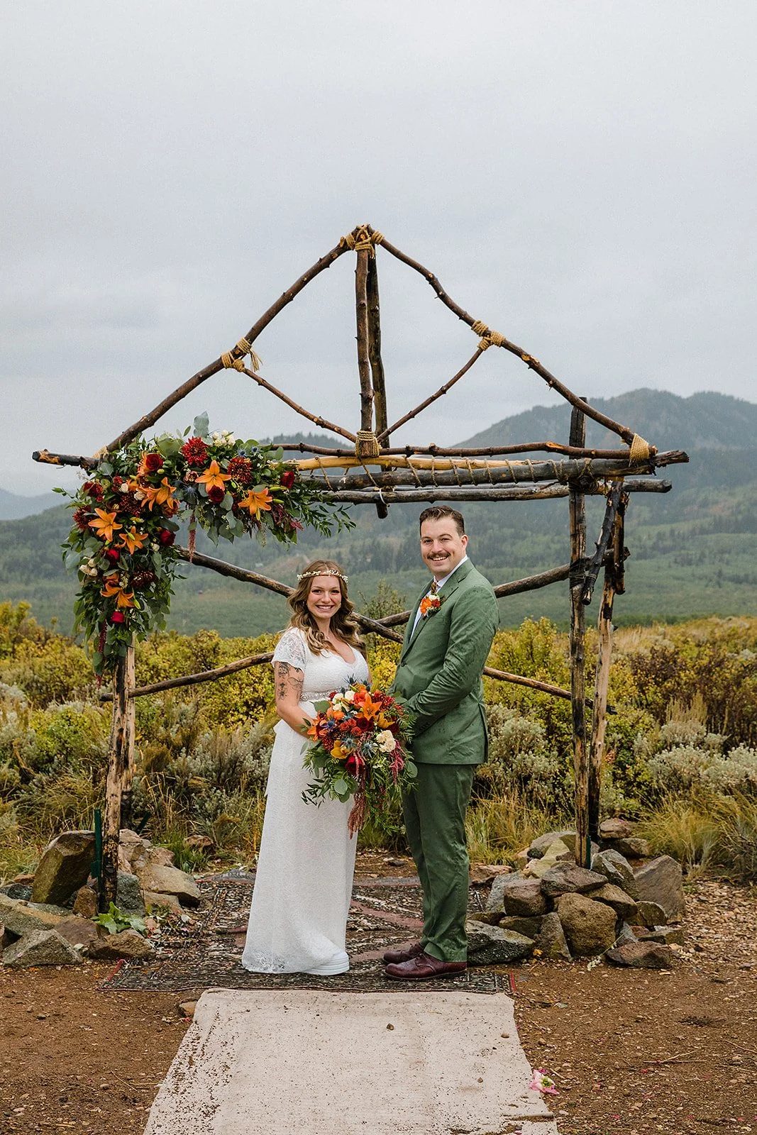 A bride and groom standing outdoors during their wedding ceremony, holding a large bouquet of flowers, in front of a rustic wooden arch decorated with flowers, with a mountainous landscape in the background.