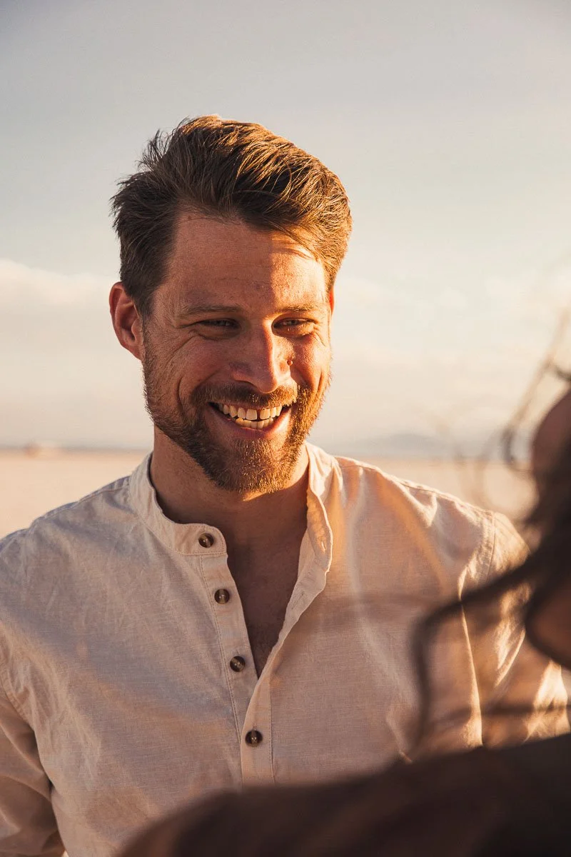 A smiling man with light brown hair and a beard, wearing a beige button-up shirt, standing outdoors at sunset.