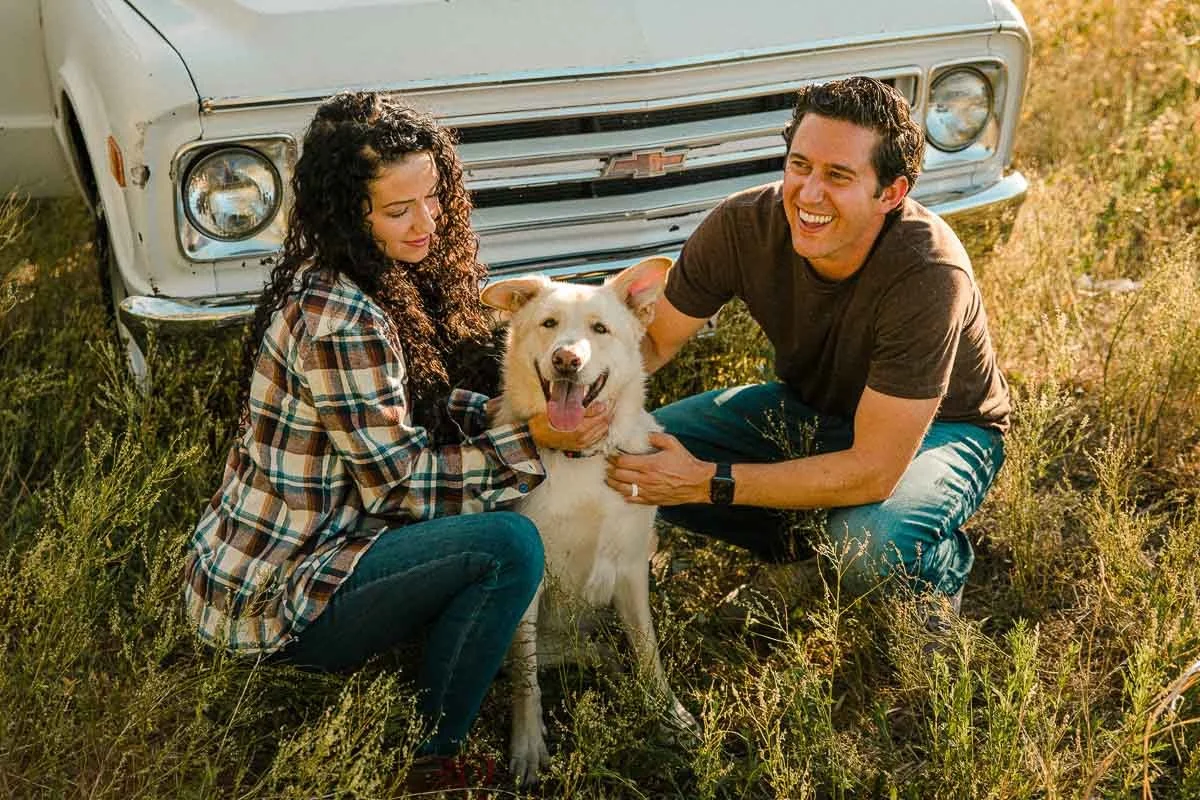 A couple sits with their dog in a grassy field next to an old white Chevrolet van, smiling and enjoying the outdoors during sunset.